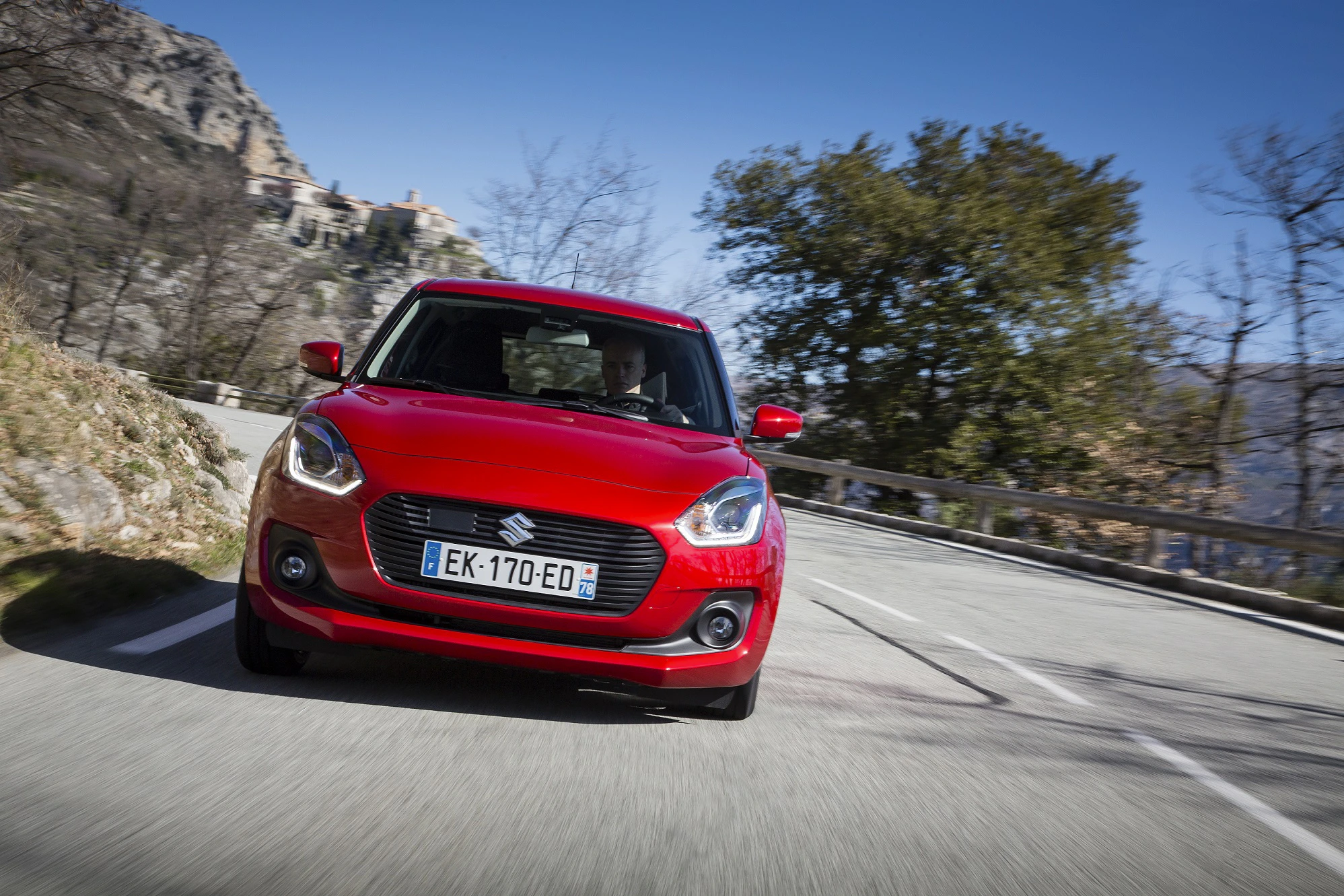 Roter Suzuki Swift fährt auf kurviger Bergstraße. Im Hintergrund Bäume und Felsen unter blauem Himmel.