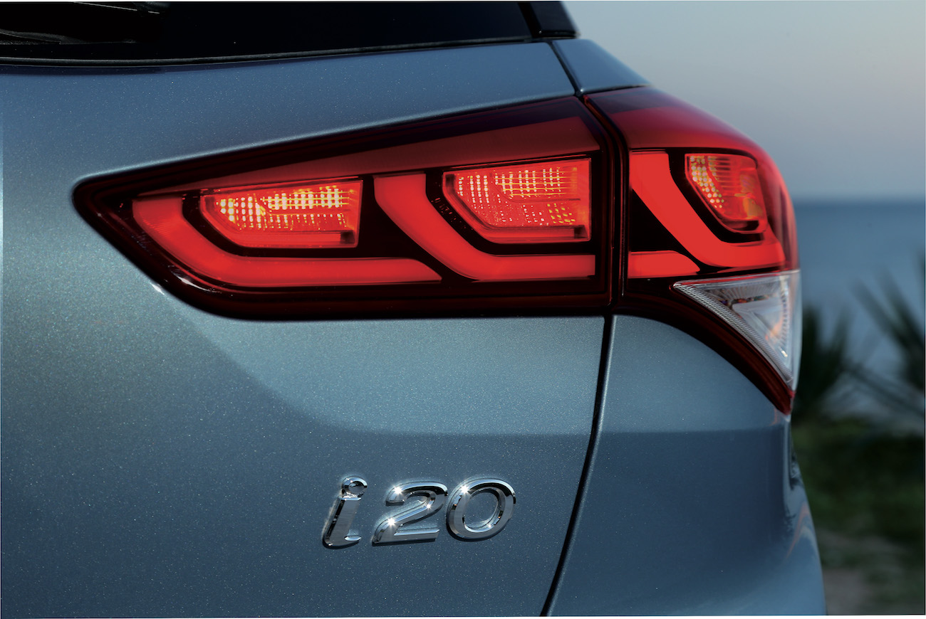 Nahaufnahme eines hellblauen Autos mit leuchtendem Rücklicht und dem Emblem "i20" am Heck. Hintergrund zeigt abendlichen Himmel.