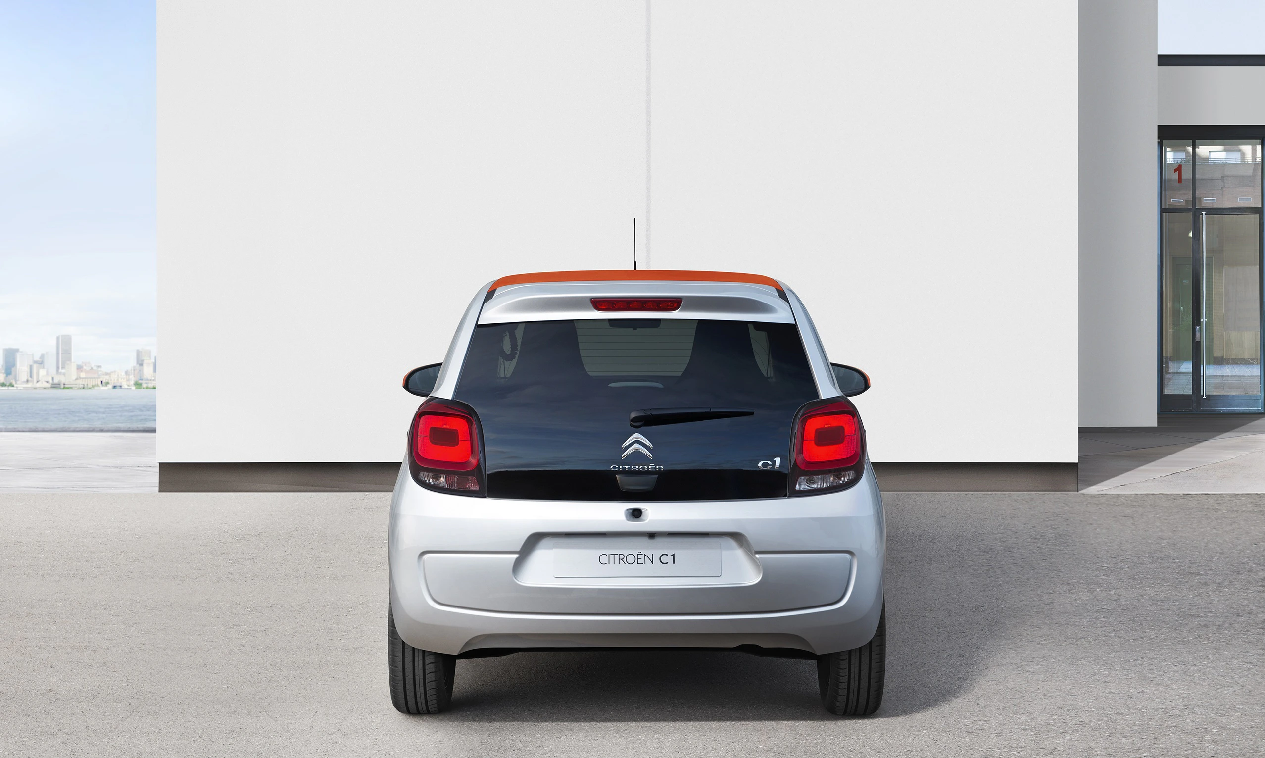 Silberner Citroën C1 mit orangenem Dach, von hinten sichtbar, vor einem modernen Gebäude geparkt.