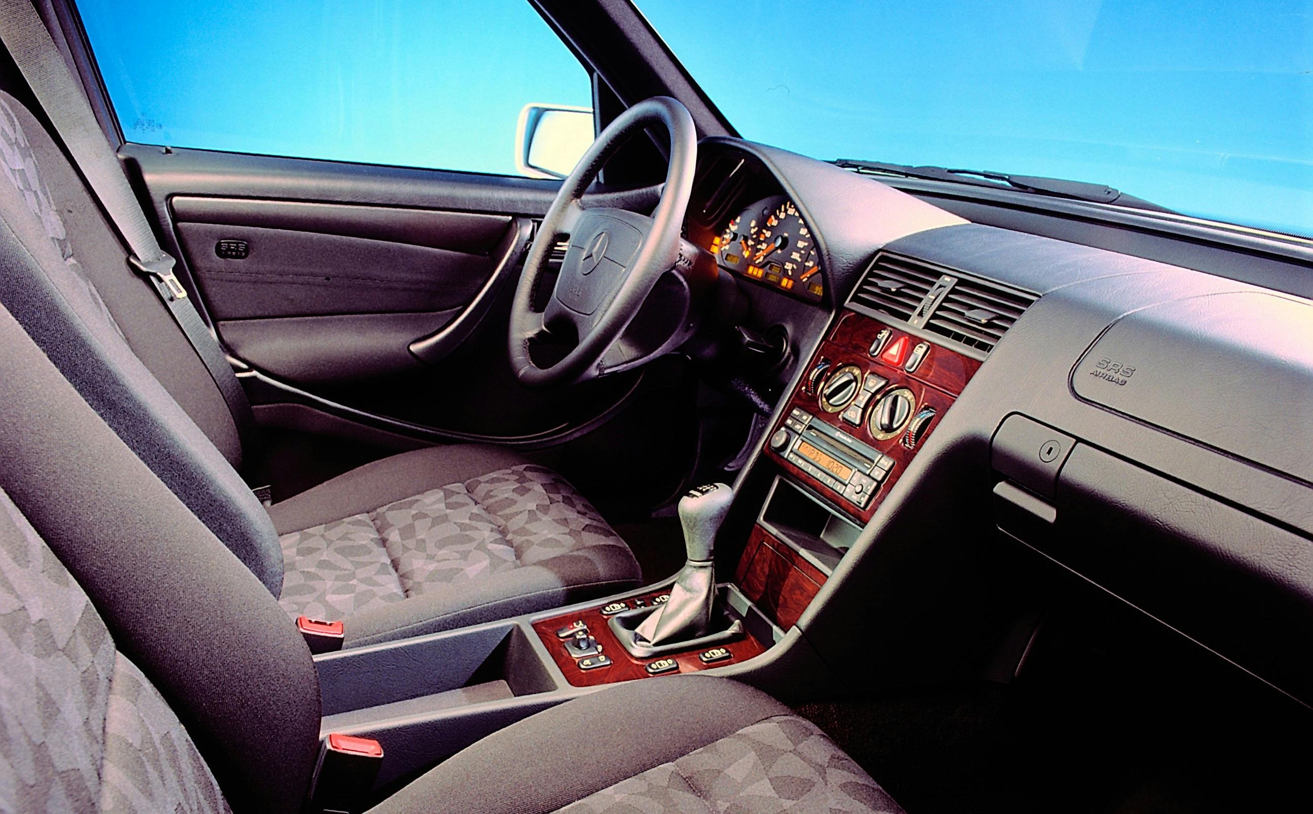 Innenraum eines Mercedes-Benz mit klassischem Armaturenbrett, Holzverkleidung, Automatikschaltung und dunkler Stoffpolsterung vor blauem Hintergrund.