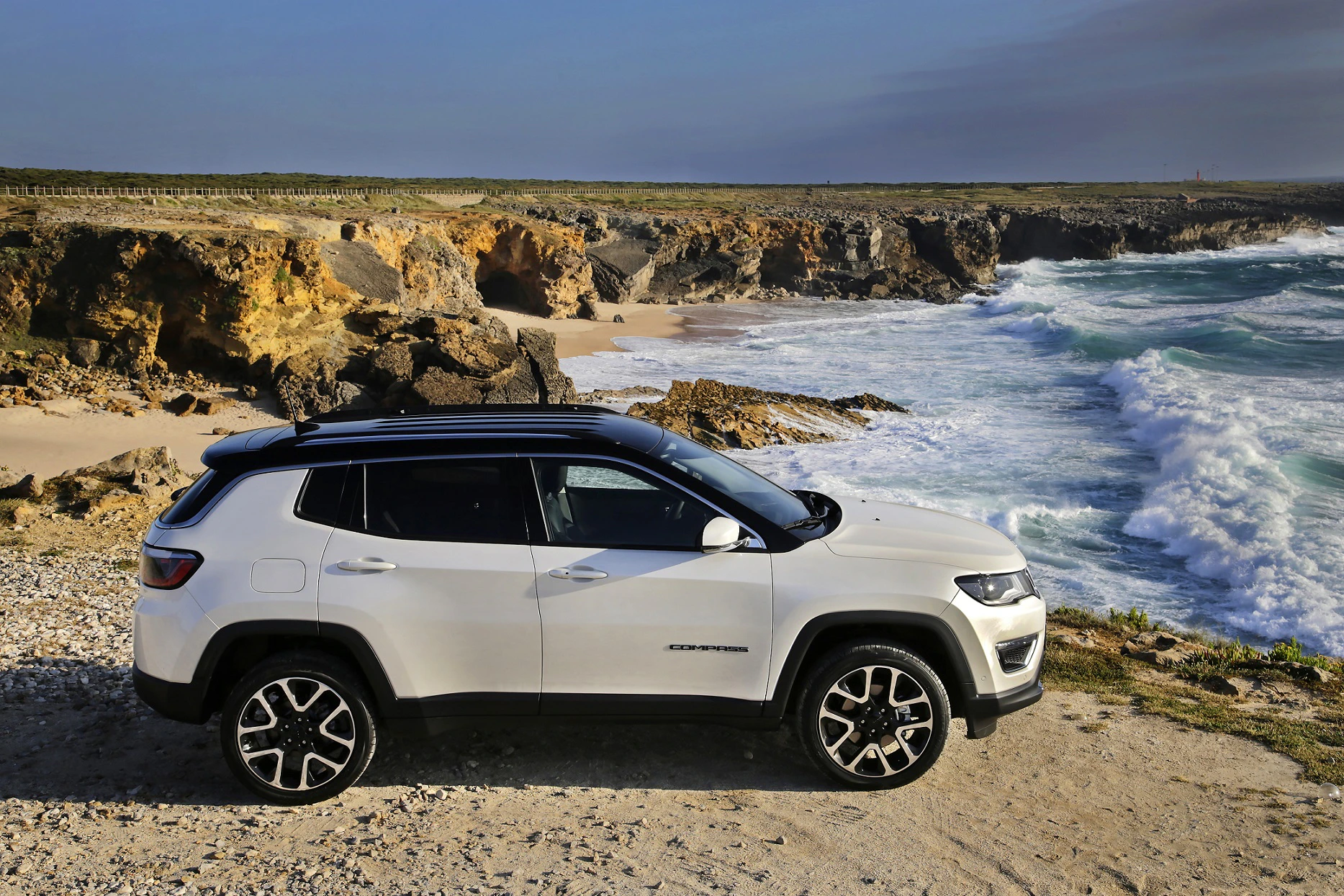 Weißer SUV parkt an einer felsigen Küste mit rauer Brandung und blauem Himmel im Hintergrund.