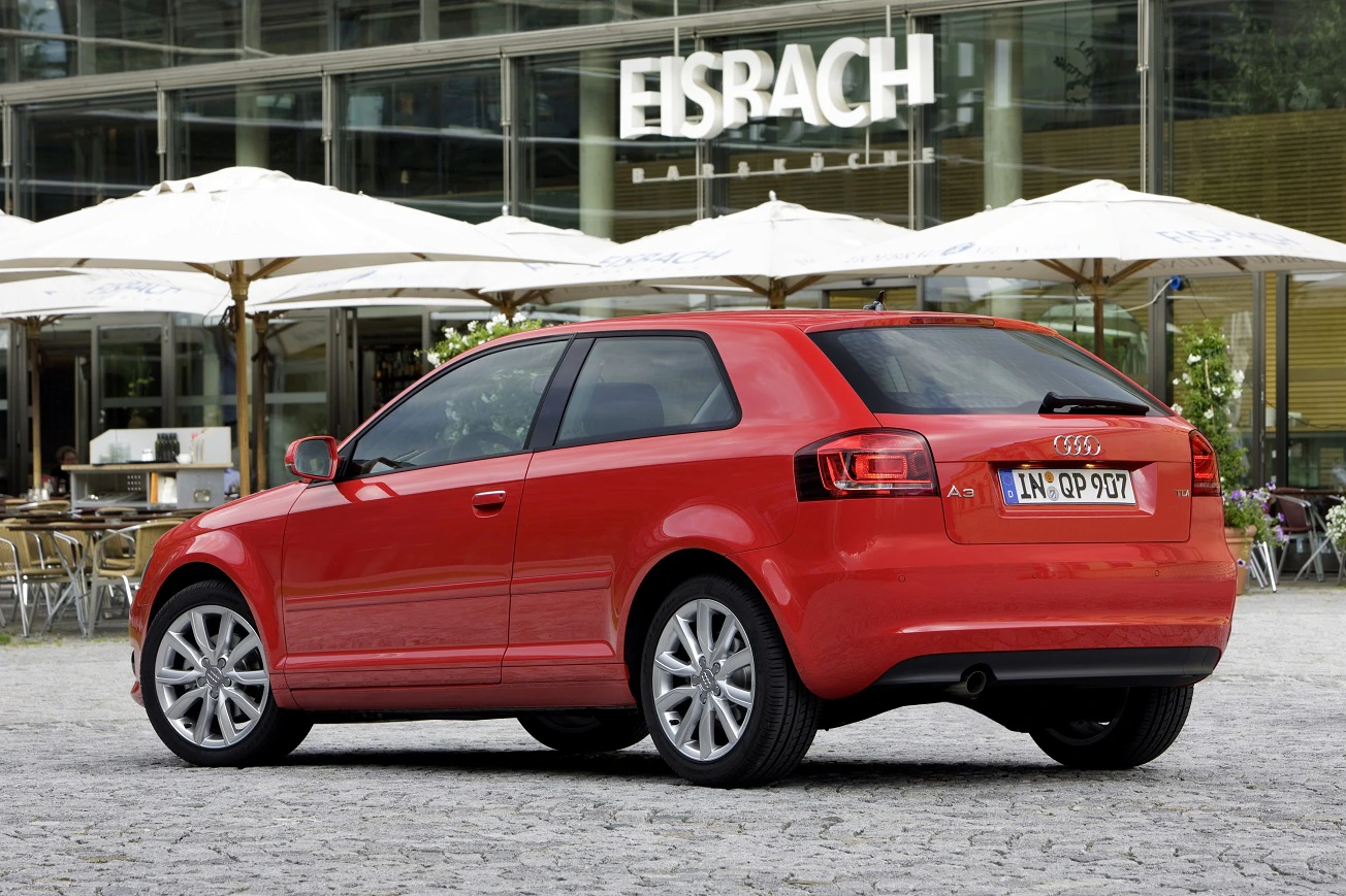 Roter Audi A3 steht auf einem gepflasterten Platz vor einem Café mit weißen Sonnenschirmen und der Aufschrift "Eisbach".