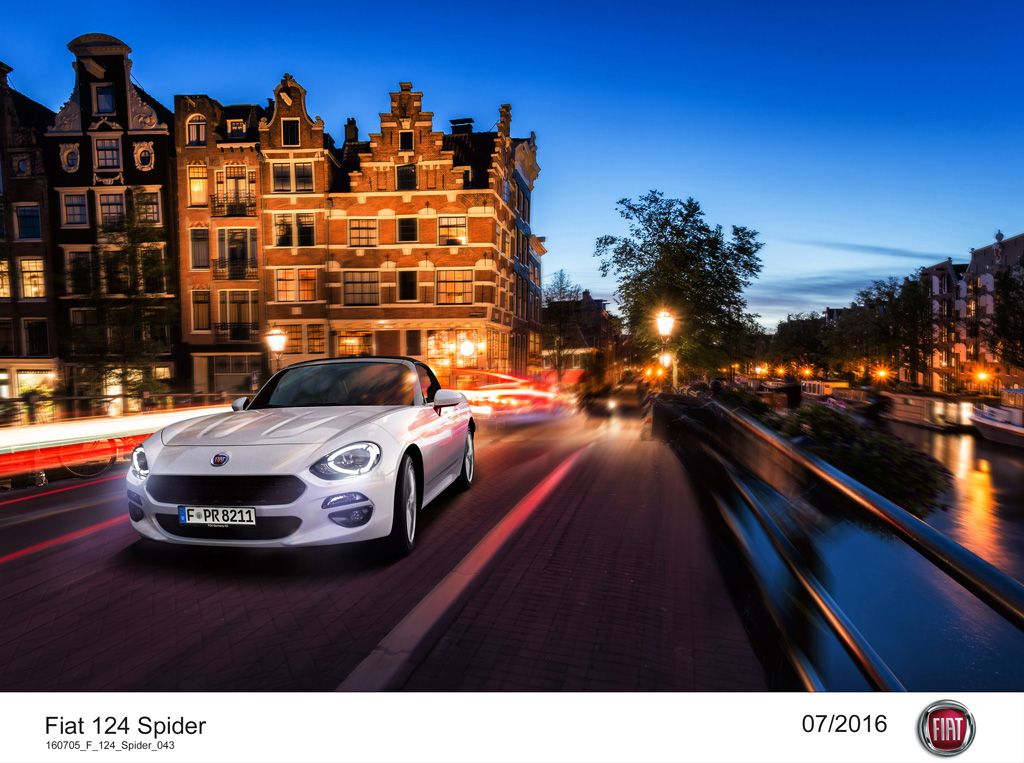 Weißer Fiat 124 Spider fährt bei Dämmerung durch eine von Kanälen gesäumte Straße mit historischen Gebäuden im Hintergrund.