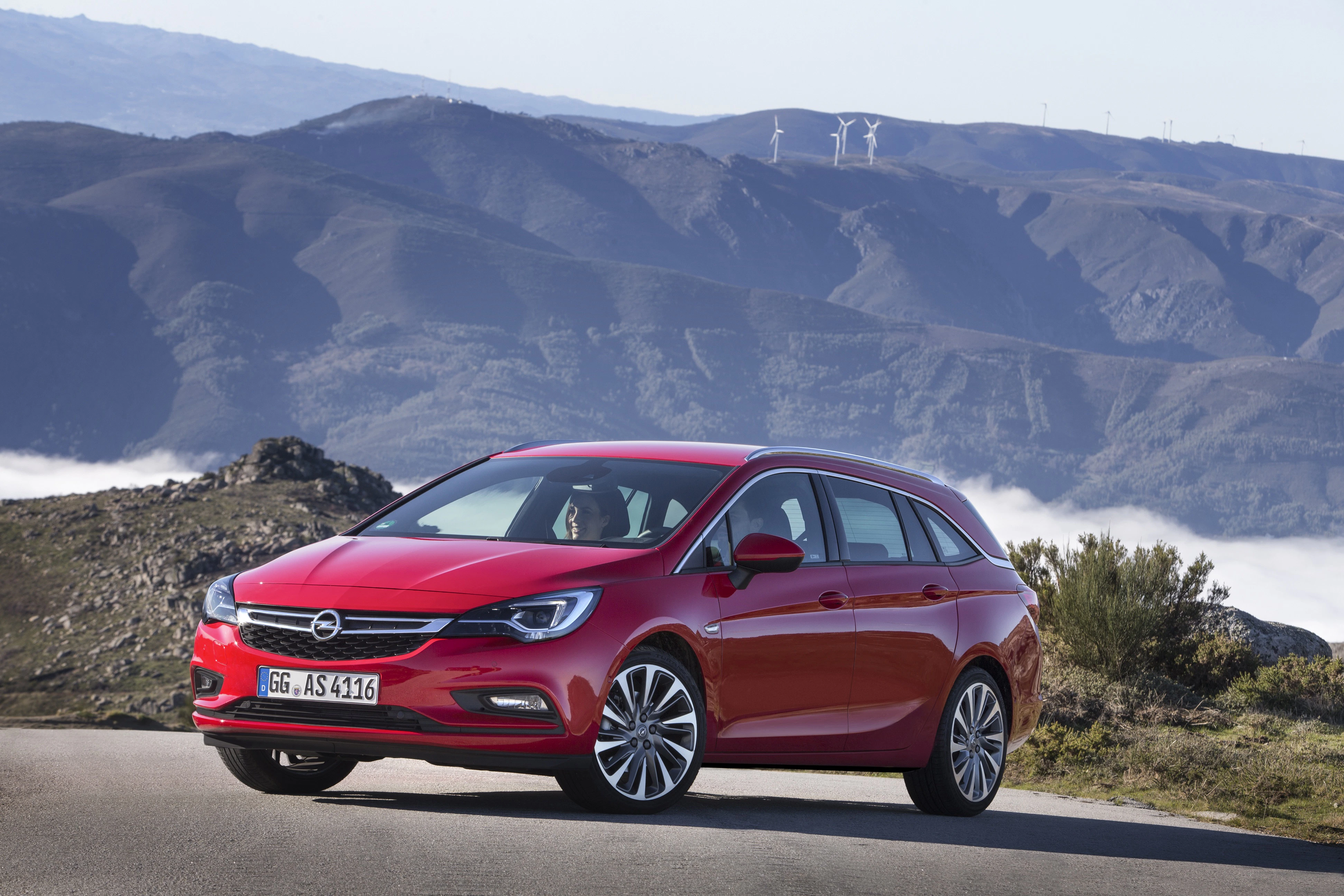 Roter Opel Astra Kombi auf einer Straße, im Hintergrund Hügel und Windräder, unter klarem Himmel.
