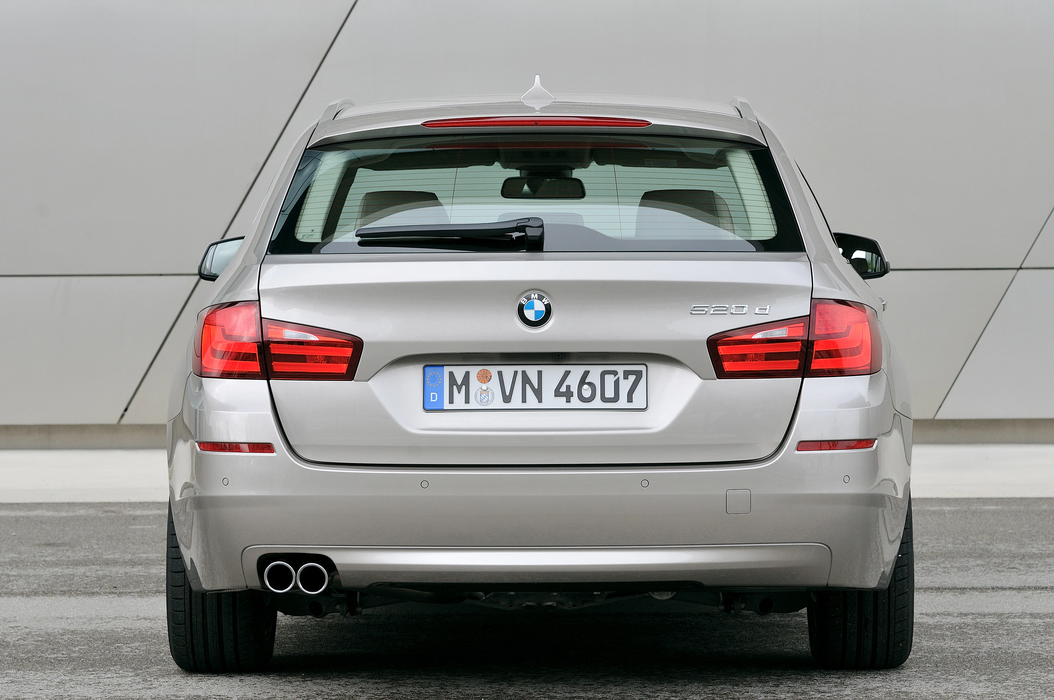 BMW 520d Touring in Silber, Rückansicht vor einer grauen Wand, duale Auspuffrohre links, Kennzeichen sichtbar.