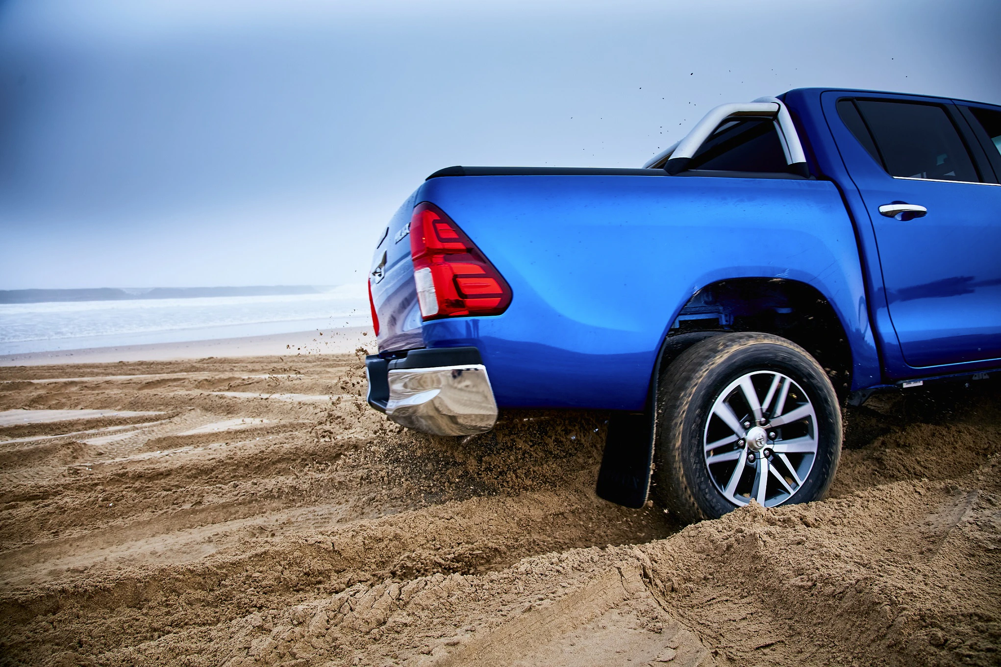 Blauer Pickup-Truck, der am Strand im Sand fährt, mit spritzendem Sand und Blick auf das Meer im Hintergrund.