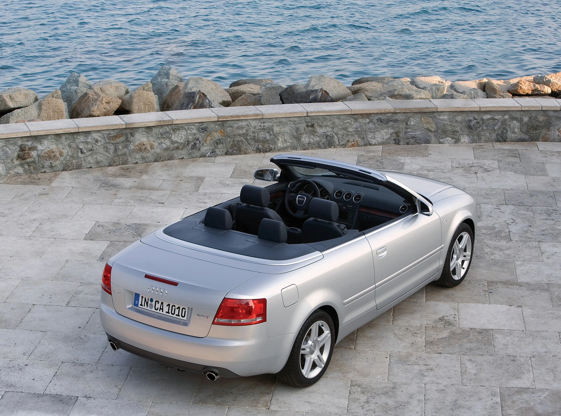 Silberner Cabrio-Pkw parkt auf einer steinernen Uferpromenade, im Hintergrund das Meer und Felsen.