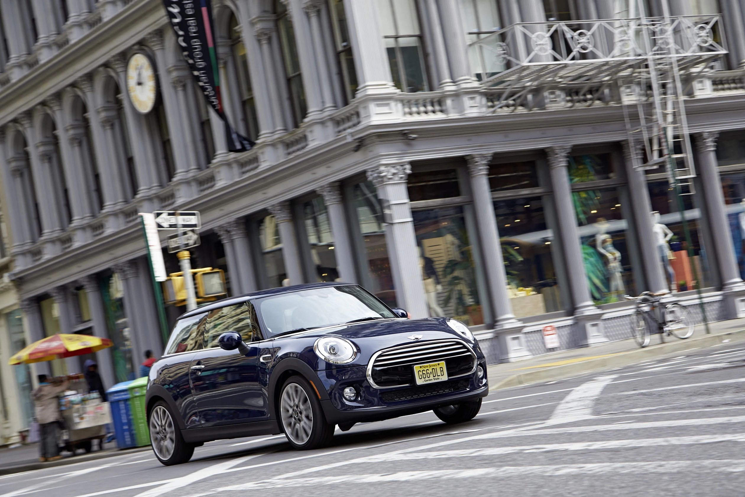 Ein blauer Mini Cooper fährt an einer Straßenecke in einer Stadt vorbei, im Hintergrund ein historisches Gebäude mit großen Schaufenstern.