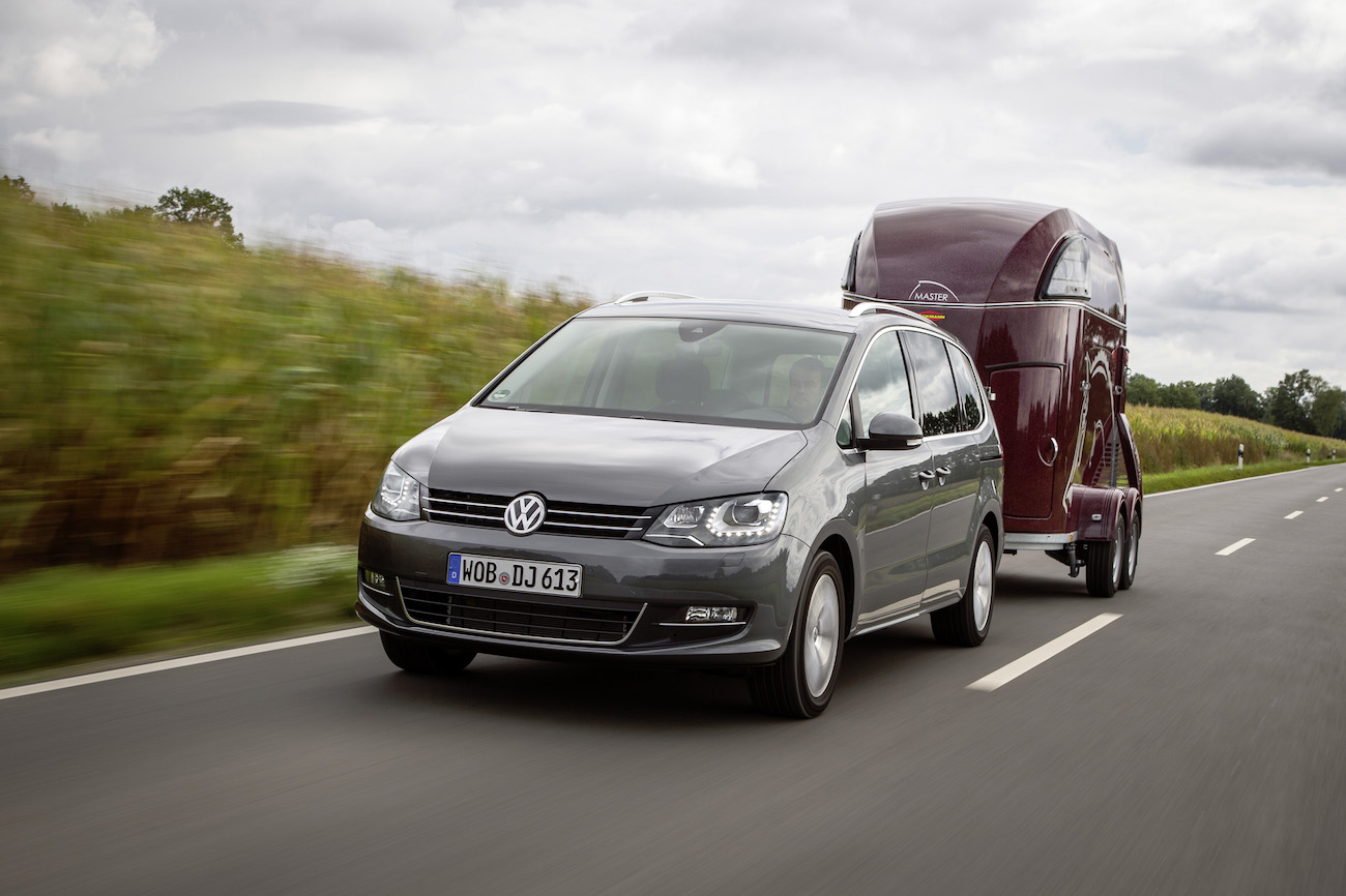 Ein grauer VW Van fährt auf einer Landstraße und zieht einen roten Pferdeanhänger bei bewölktem Himmel.