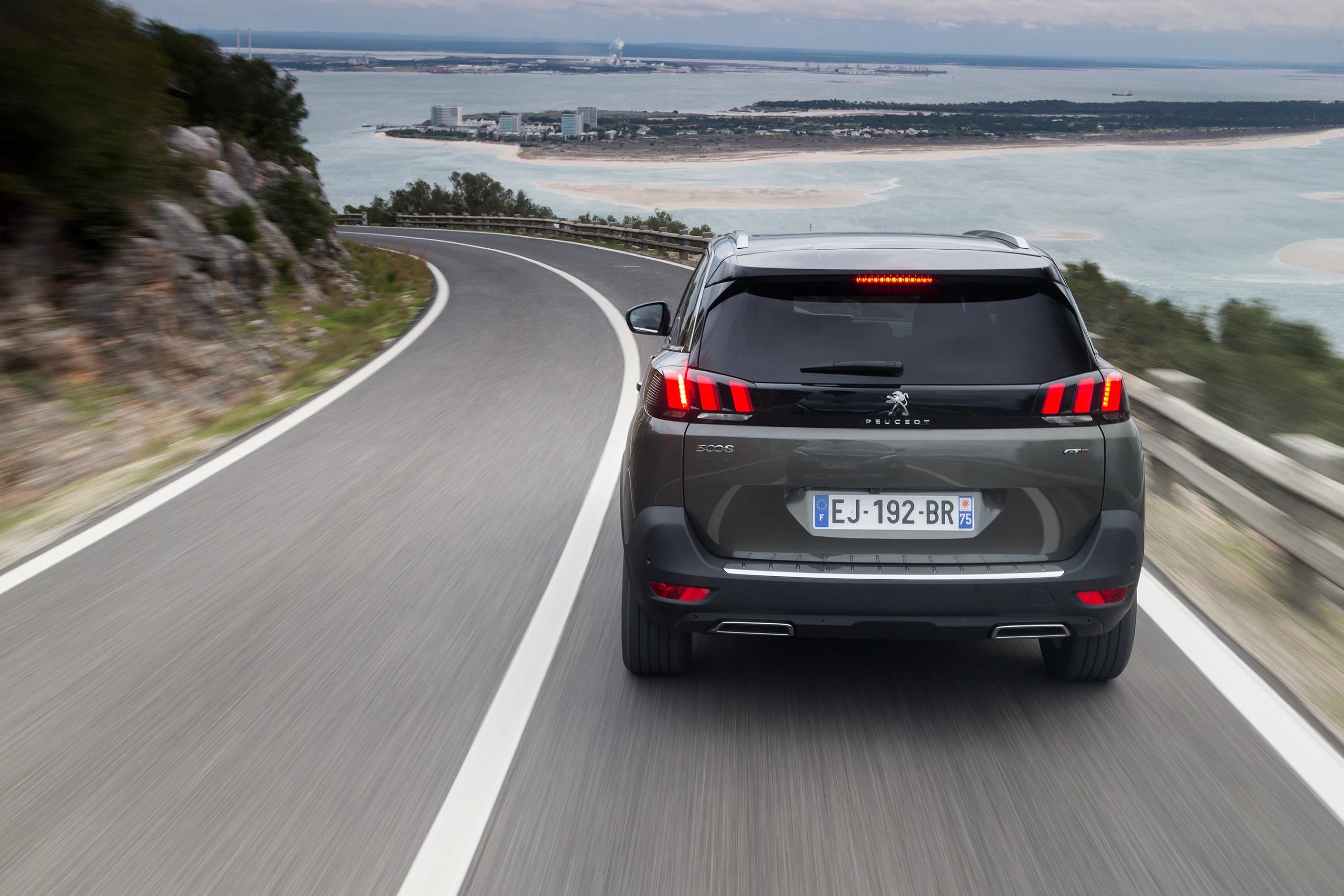 Ein schwarzer SUV fährt auf einer kurvenreichen Küstenstraße mit Meeresblick im Hintergrund.