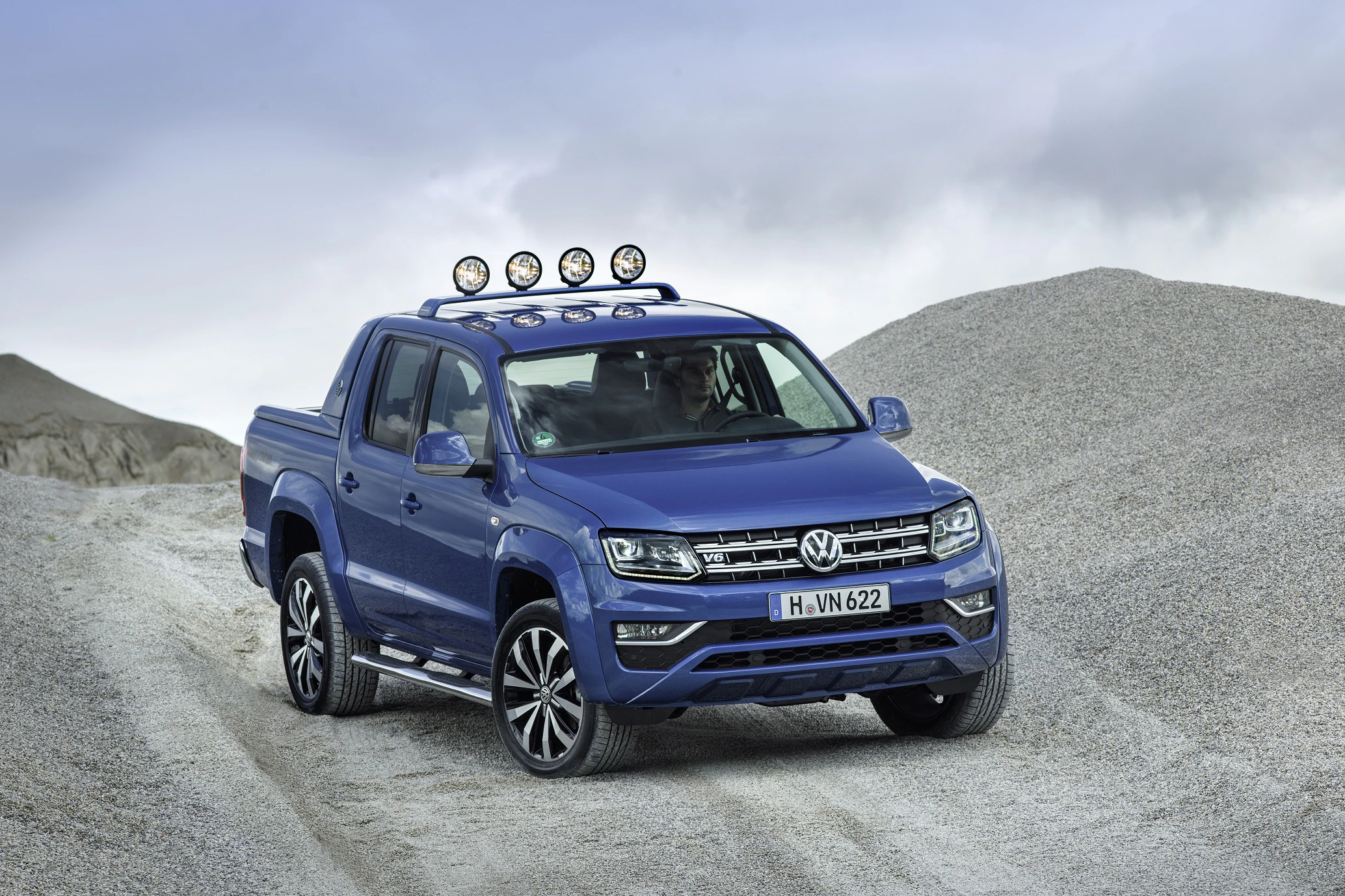 Blauer Volkswagen Amarok mit Zusatzscheinwerfern fährt über eine Kiesfläche. Dynamische Kulisse mit bewölktem Himmel im Hintergrund.
