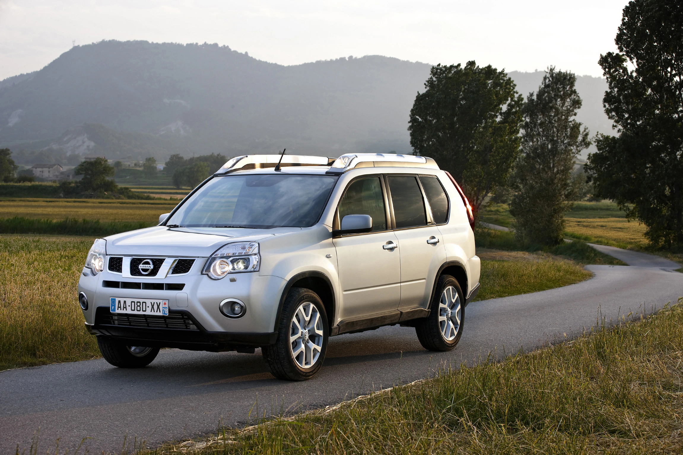 Silberner SUV auf einer Landstraße, umgeben von Wiesen und Hügeln im Hintergrund. Die Sonne steht tief am Himmel.
