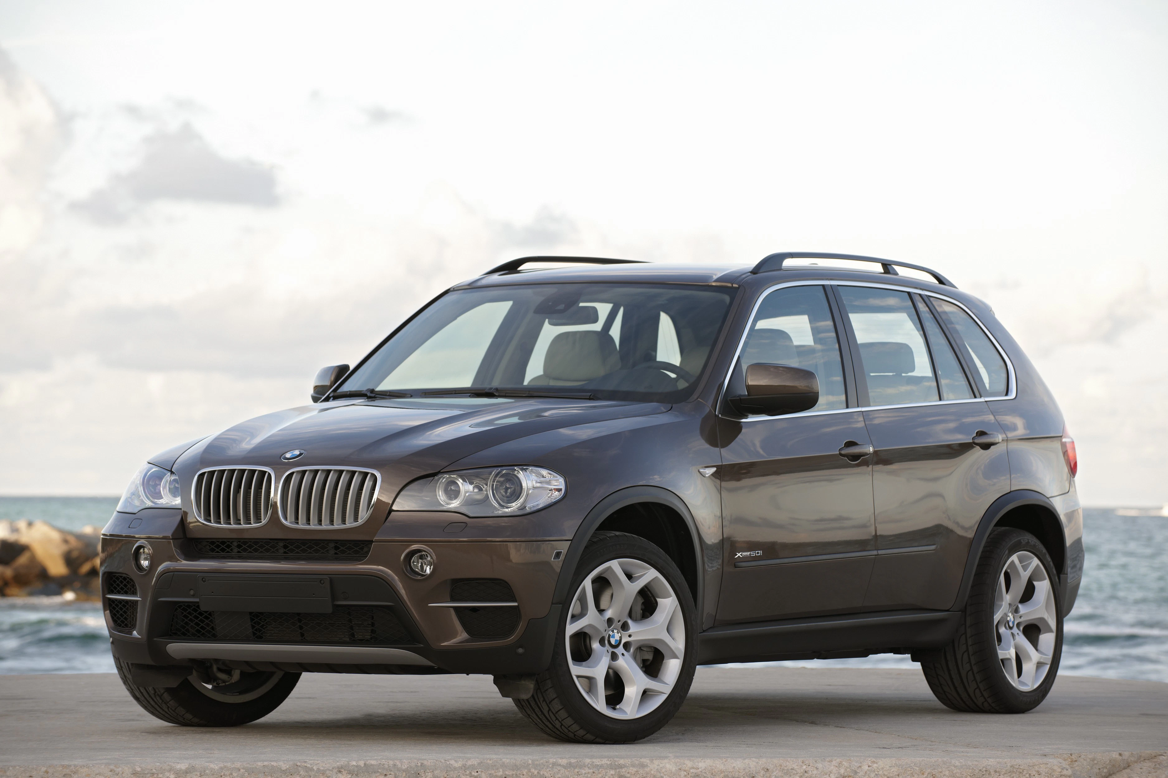 Brauner BMW X5 SUV steht auf einem Parkplatz am Meer, leicht bewölkter Himmel im Hintergrund.