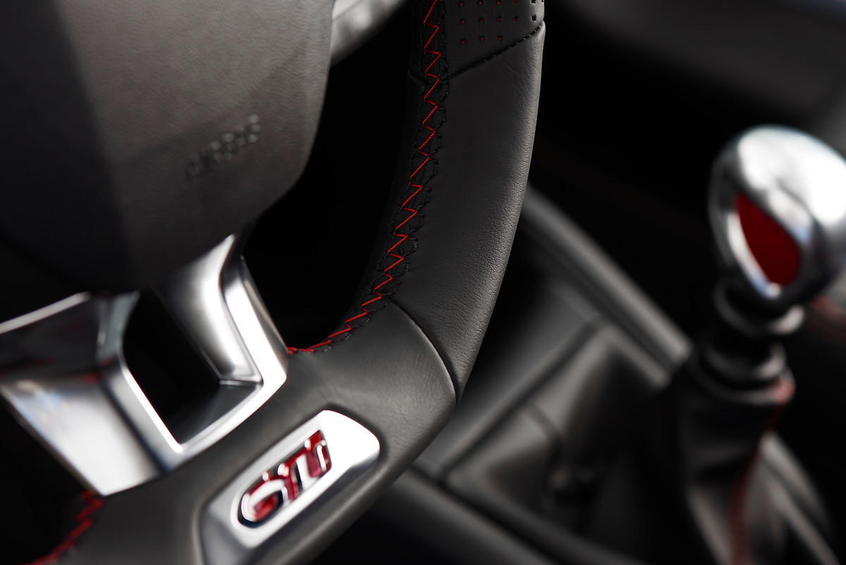 Nahaufnahme eines Lenkrads mit GTI-Emblem und roter Naht. Im Hintergrund ist ein Schalthebel sichtbar.