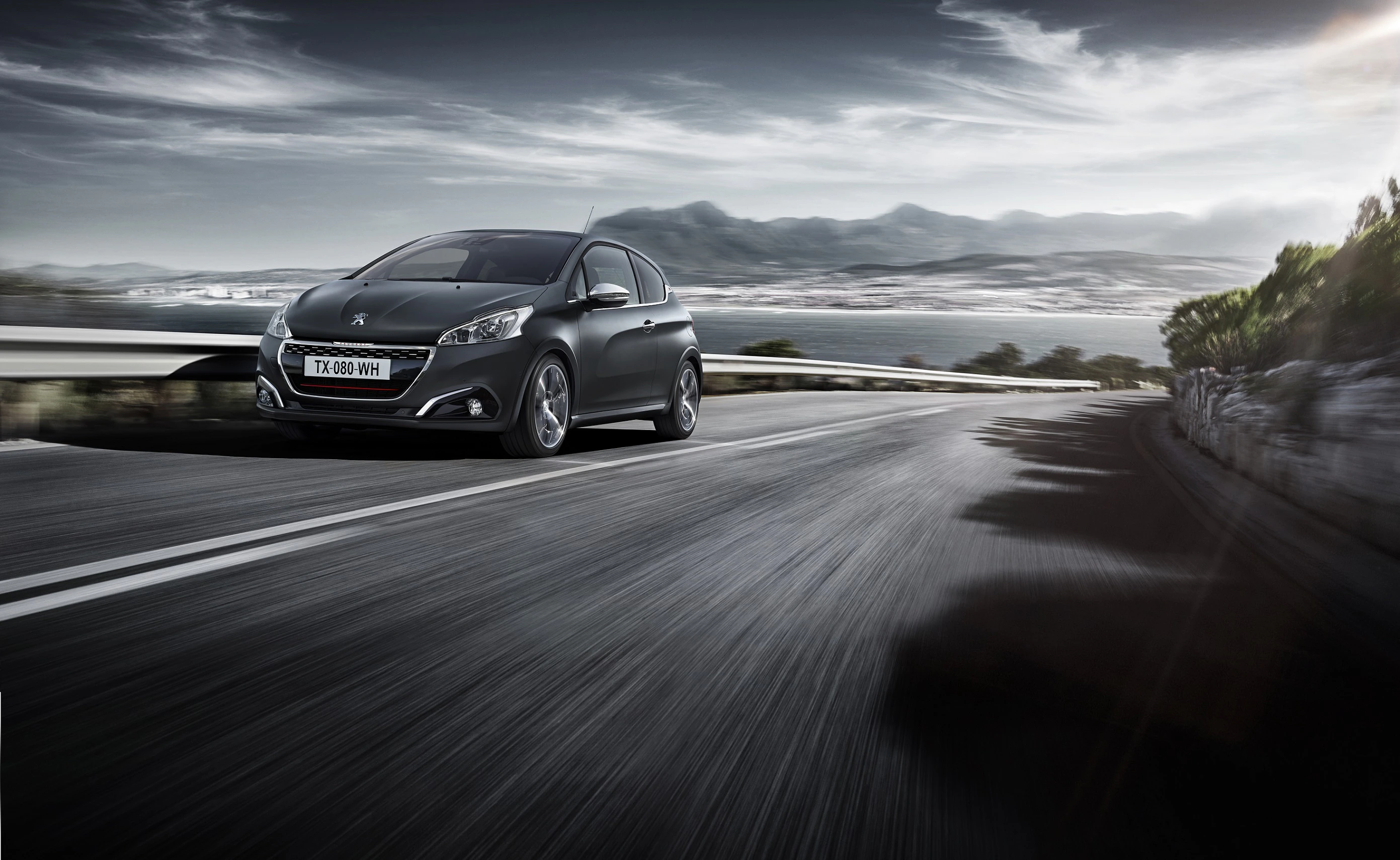 Ein grauer Peugeot fährt dynamisch auf einer kurvigen Küstenstraße, mit im Hintergrund Bergen und einem bewölkten Himmel.