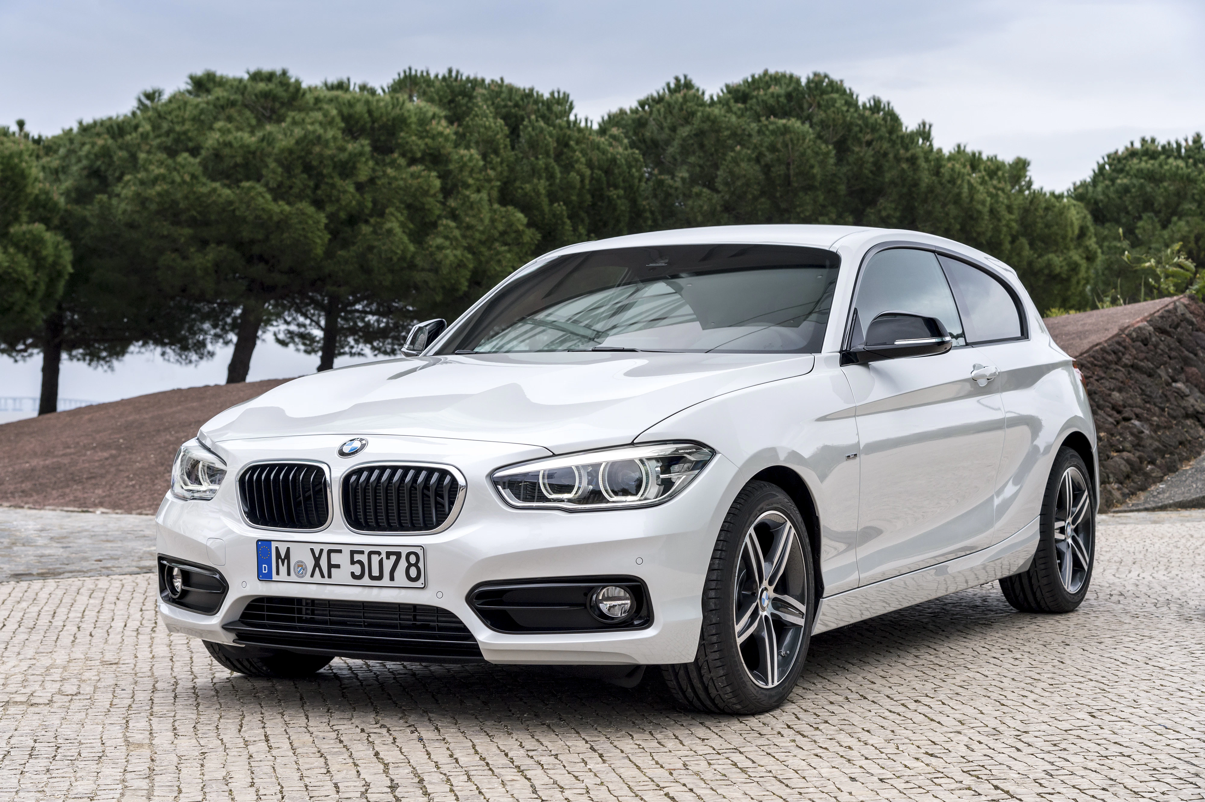 Weißer BMW 1er F20 steht auf gepflastertem Weg, umgeben von Bäumen und Natur im Hintergrund.