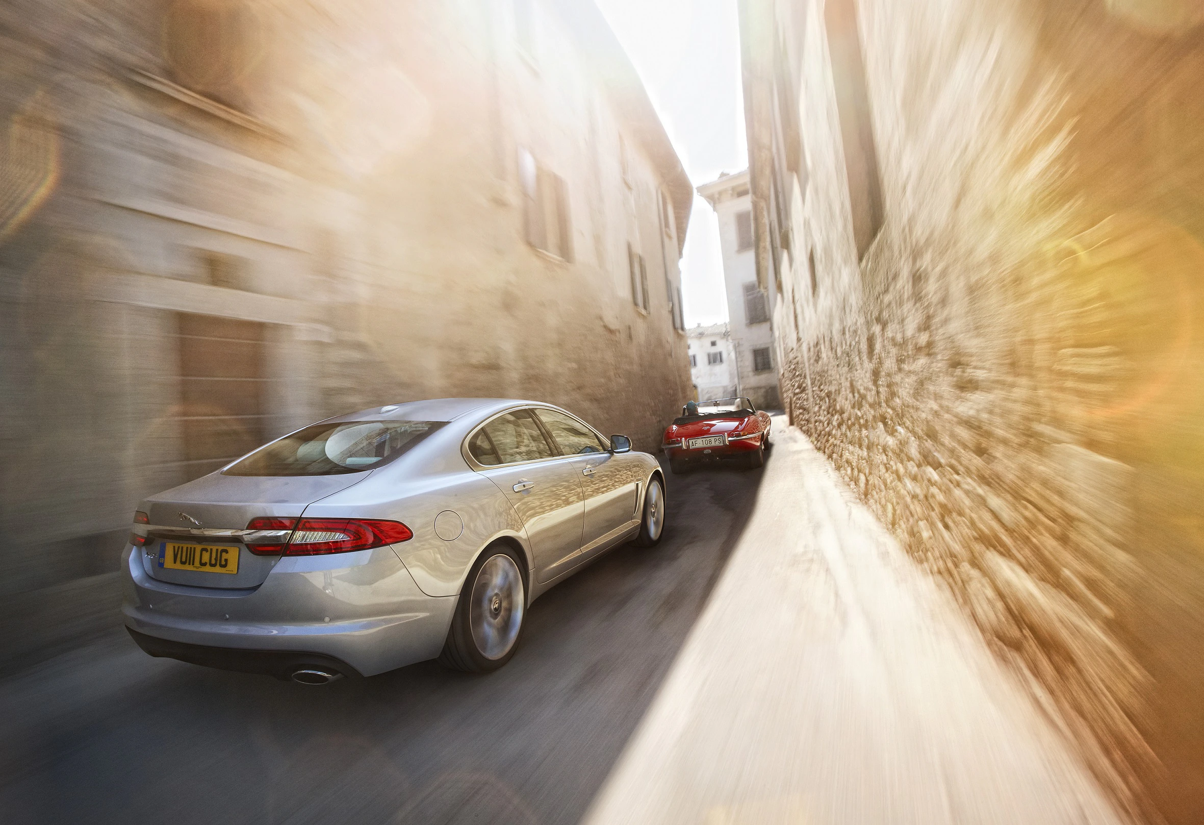 Zwei Autos fahren schnell durch enge, sonnendurchflutete Gasse einer alten Stadt mit hohen Steinmauern. Lichtstrahlen verstärken die Dynamik.
