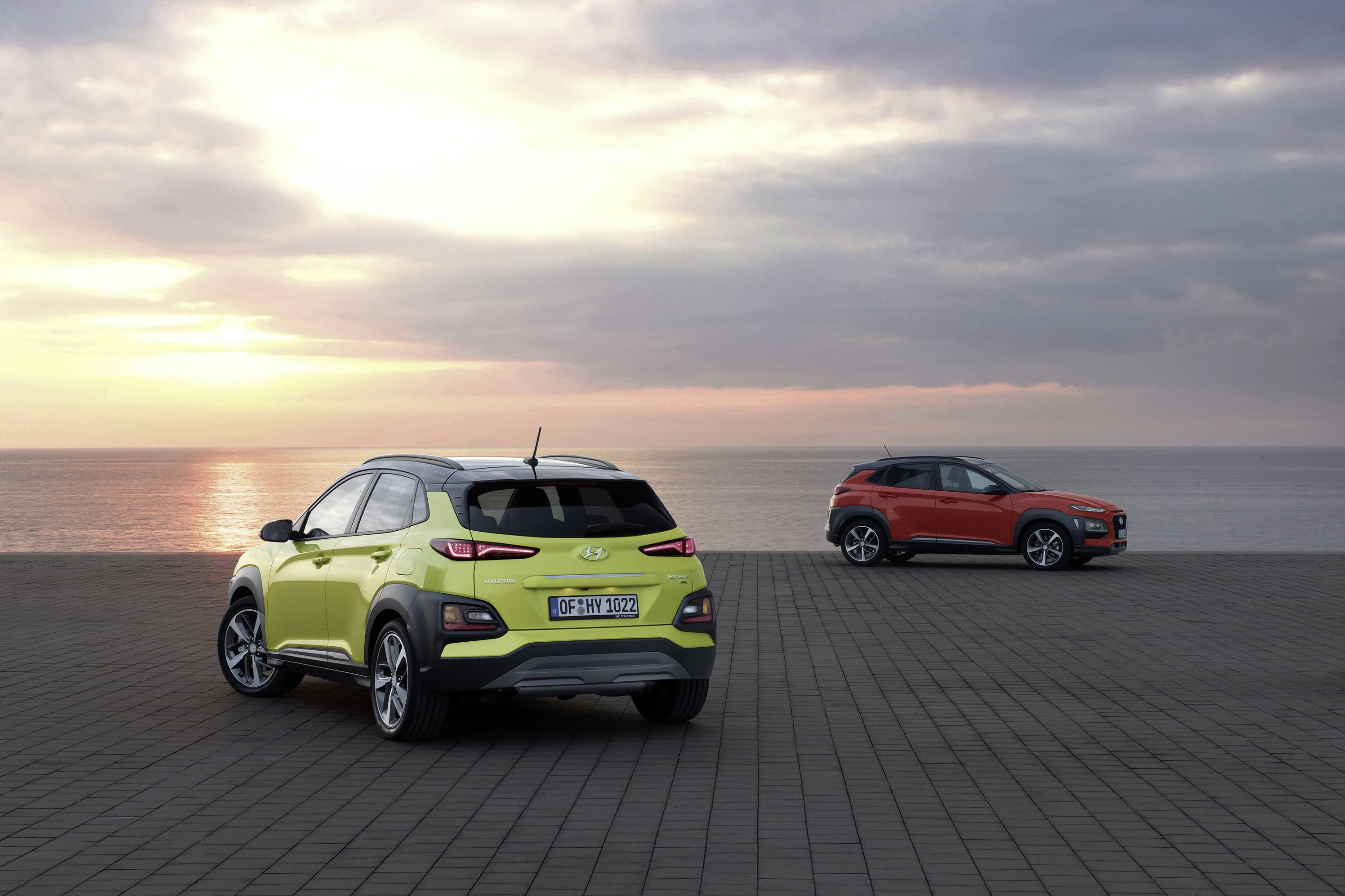 Zwei farbenfrohe Autos am Meer bei Sonnenuntergang, eines grün, das andere rot. Wolkenverhangener Himmel über ruhigem Wasser.