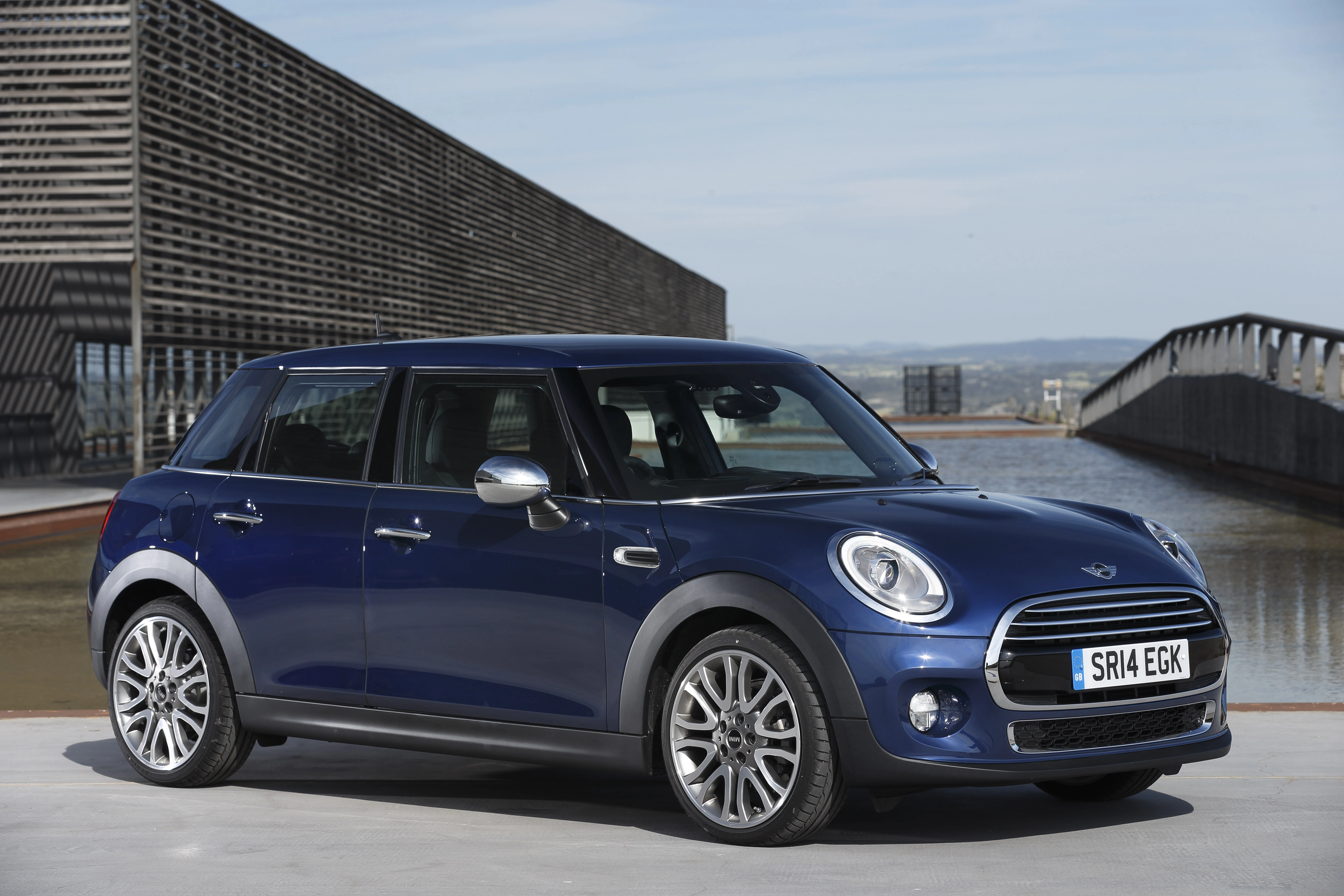 Blauer Mini Cooper Fünftürer steht vor einem modernen Gebäude mit Wasser im Hintergrund. Klarer Himmel und sonniges Wetter.