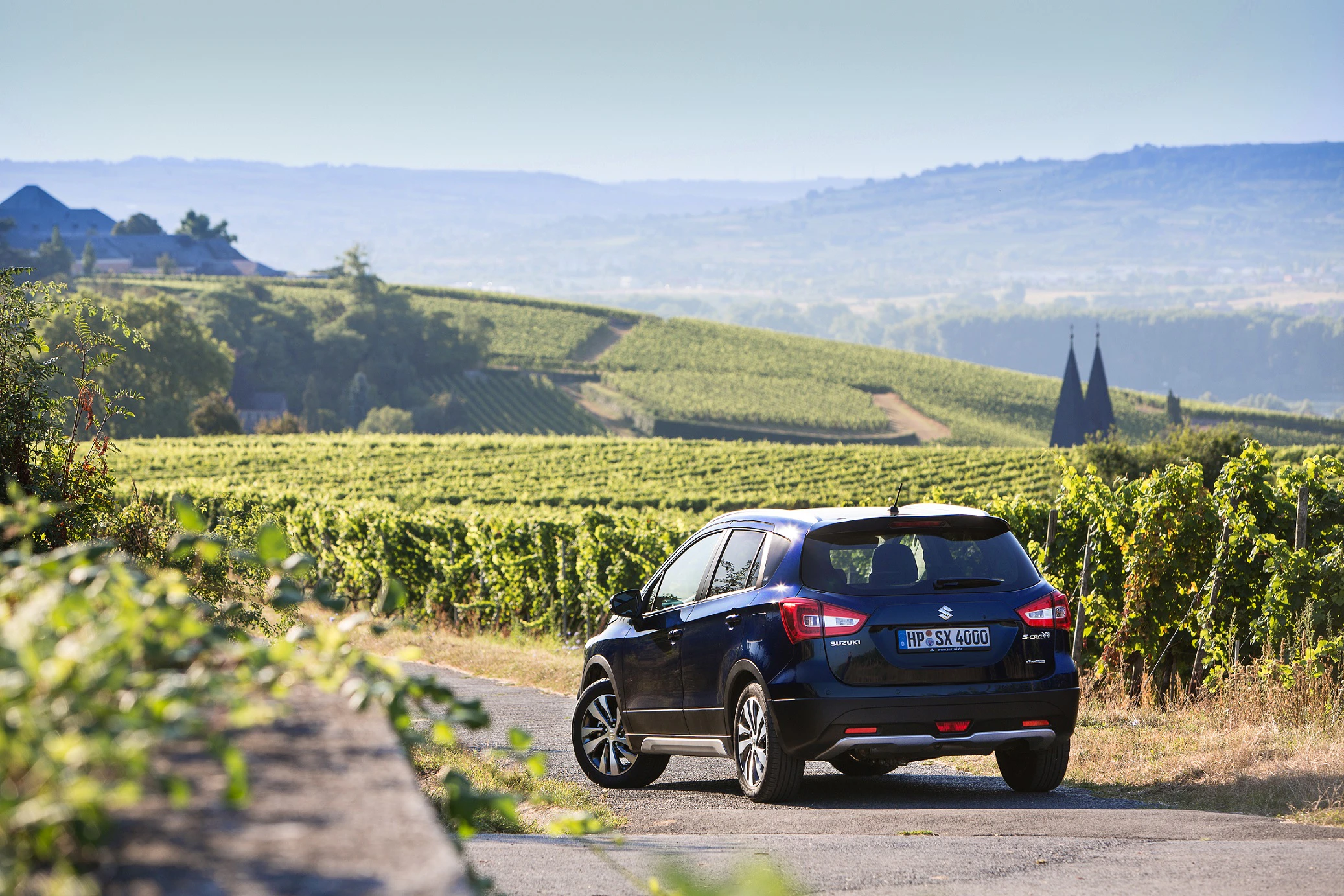 Ein blauer SUV fährt auf einer Straße durch eine malerische Weinberglandschaft bei klarem Himmel. Im Hintergrund sind Hügel sichtbar.