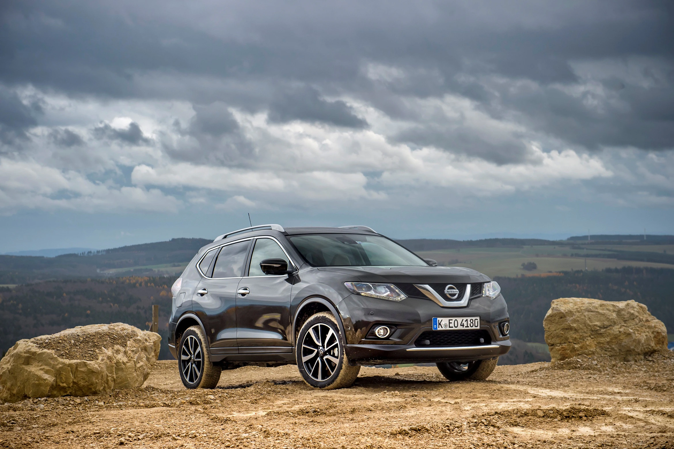 SUV auf einem Hügel mit steiniger Landschaft im Vordergrund, bedecktem Himmel und weitem Tal im Hintergrund.