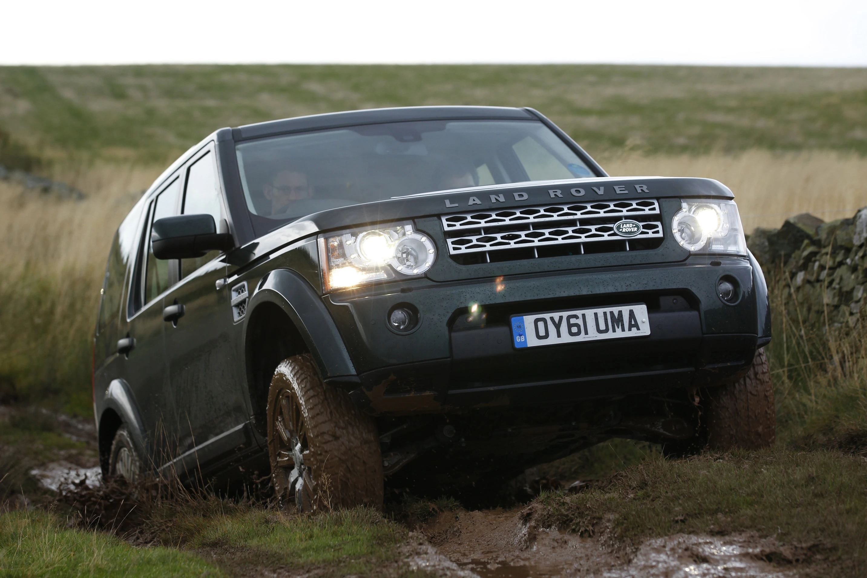 Ein schwarzer Land Rover fährt über matschiges Gelände in einer ländlichen Umgebung. Im Hintergrund sind Gräser und ein Steingebäude zu sehen.