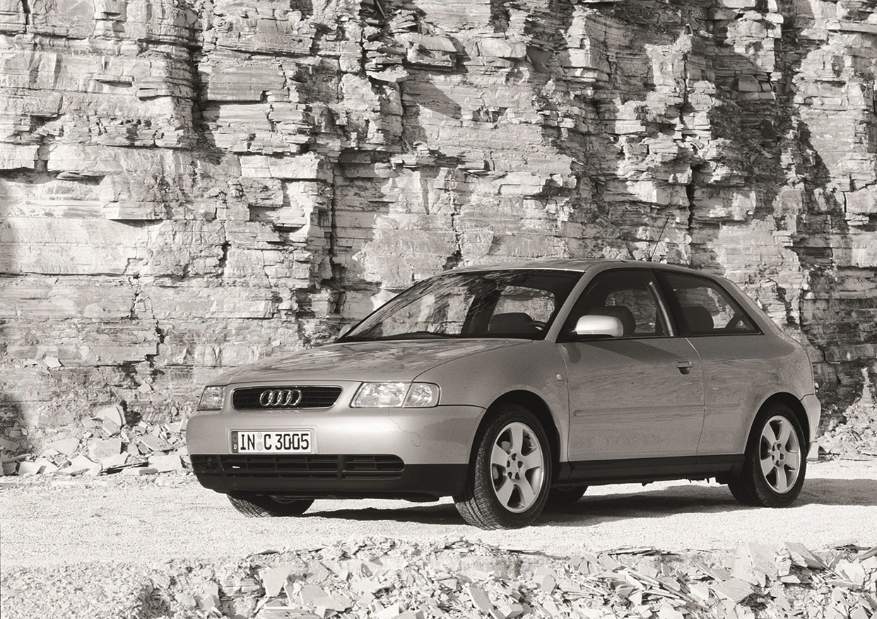 Ein silberner Audi vor einer Felswand, parkend auf Schotterboden. Das Auto ist ein älteres Modell mit einer charakteristischen, kompakten Form.