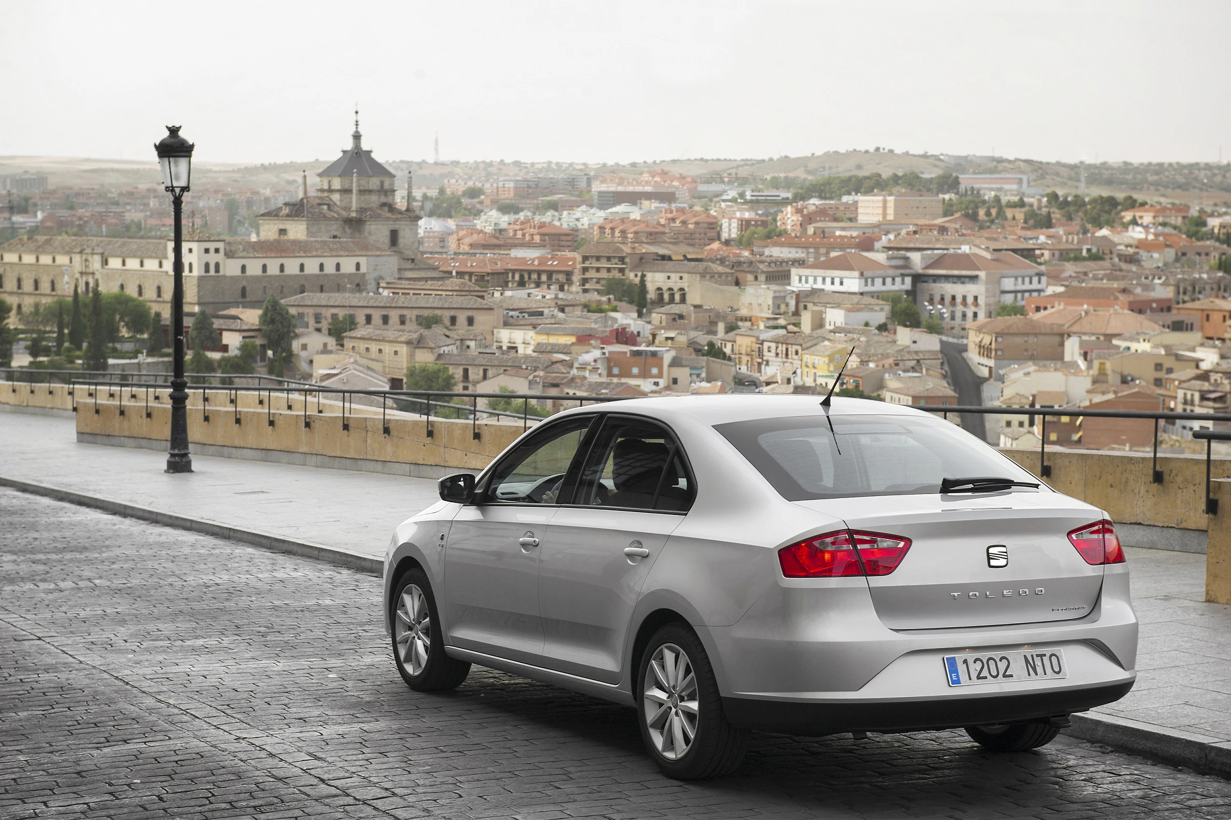 Silberner SEAT Toledo auf einer gepflasterten Straße mit Panoramablick auf eine historische Stadt im Hintergrund.