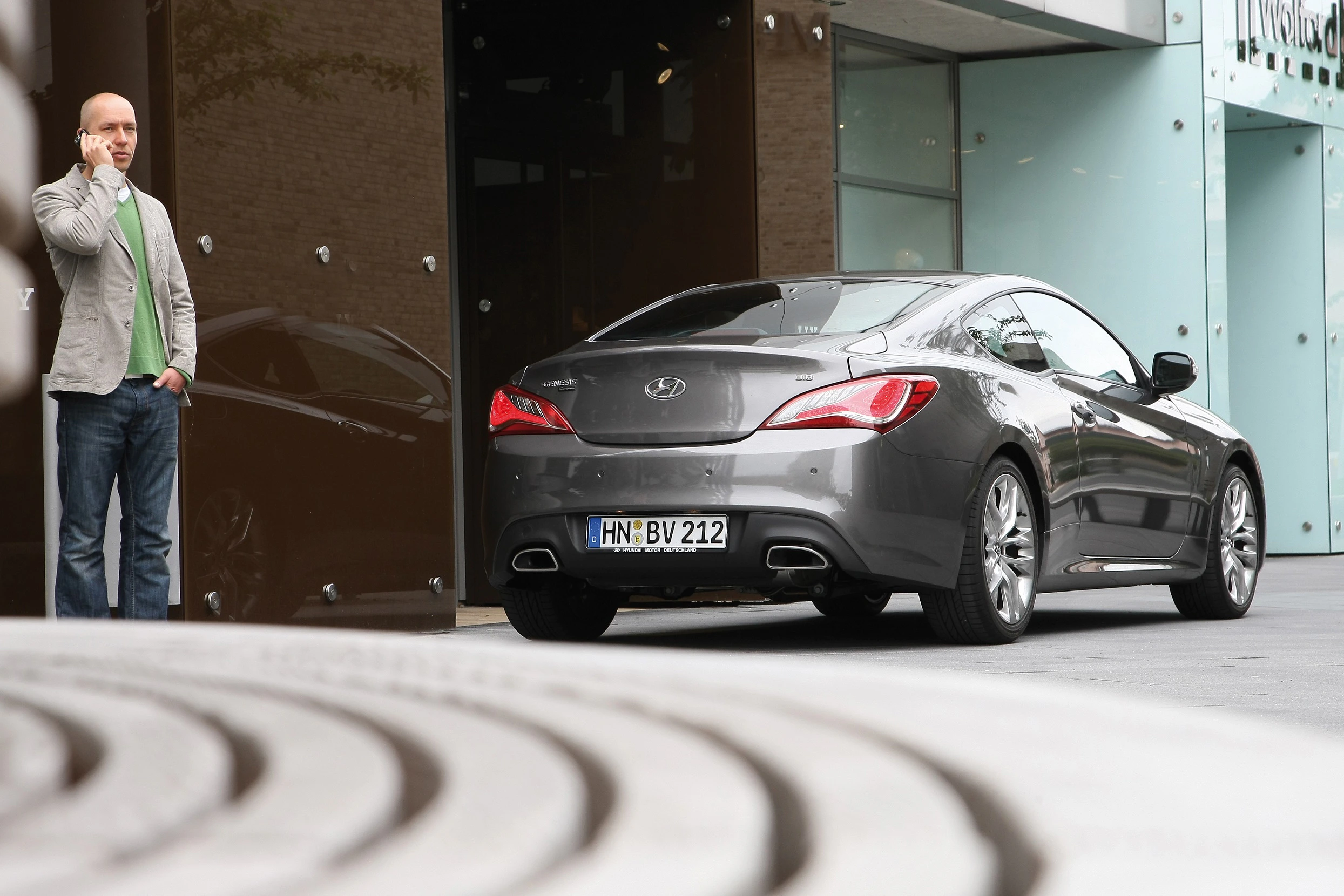 Ein Mann telefoniert neben einem grauen Coupé, das vor einem modernen Gebäude geparkt ist.