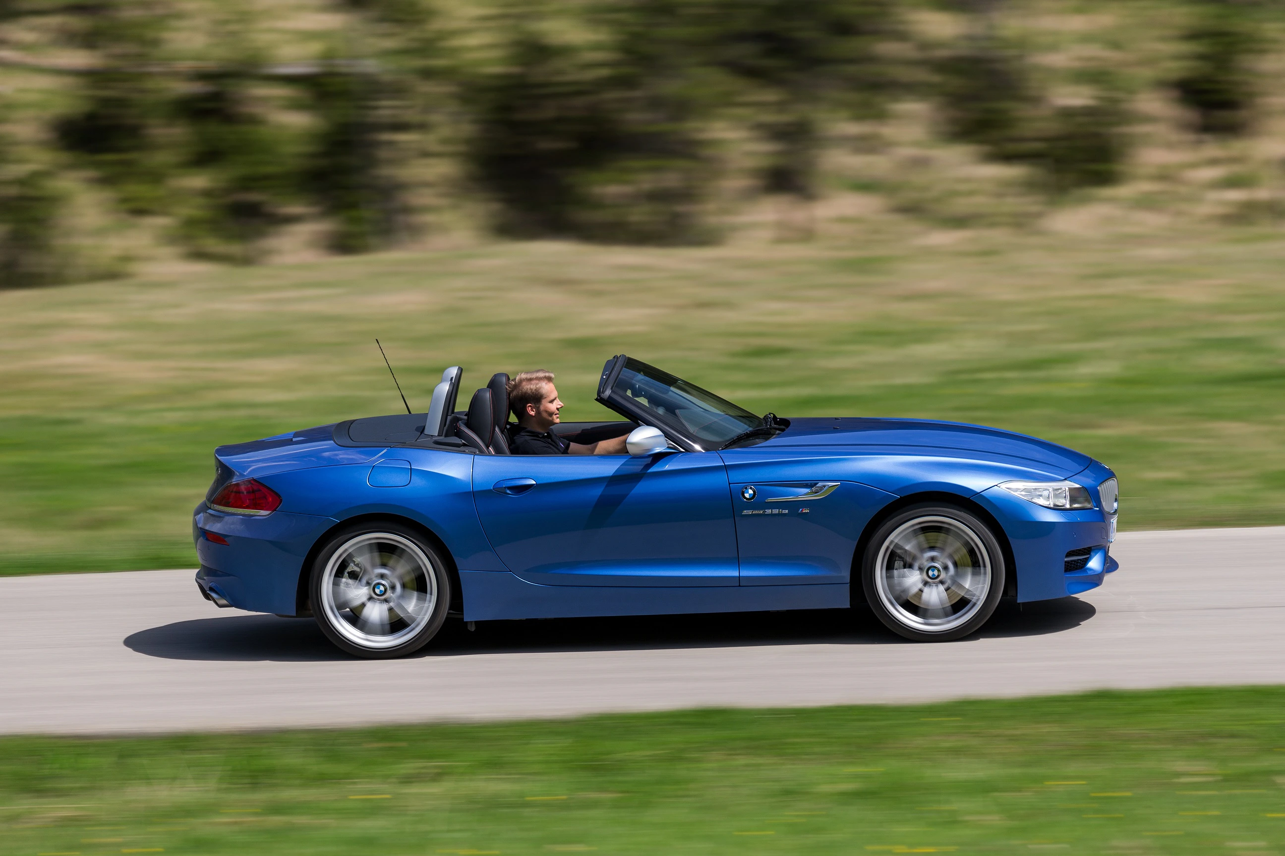 Ein blauer Sportwagen-Cabrio fährt auf einer Landstraße. Der Fahrer hat das Verdeck geöffnet und genießt die Fahrt.