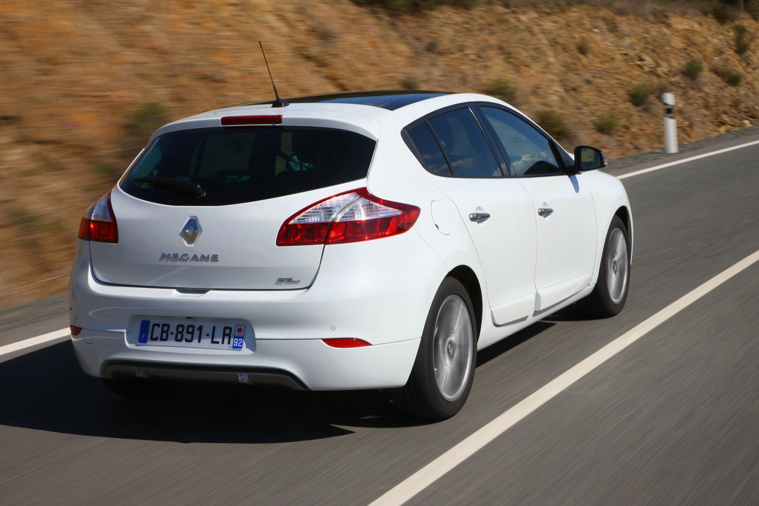 Weißer Renault Mégane fährt auf einer Landstraße; Hintergrund mit braunen Hügeln und blauem Himmel.