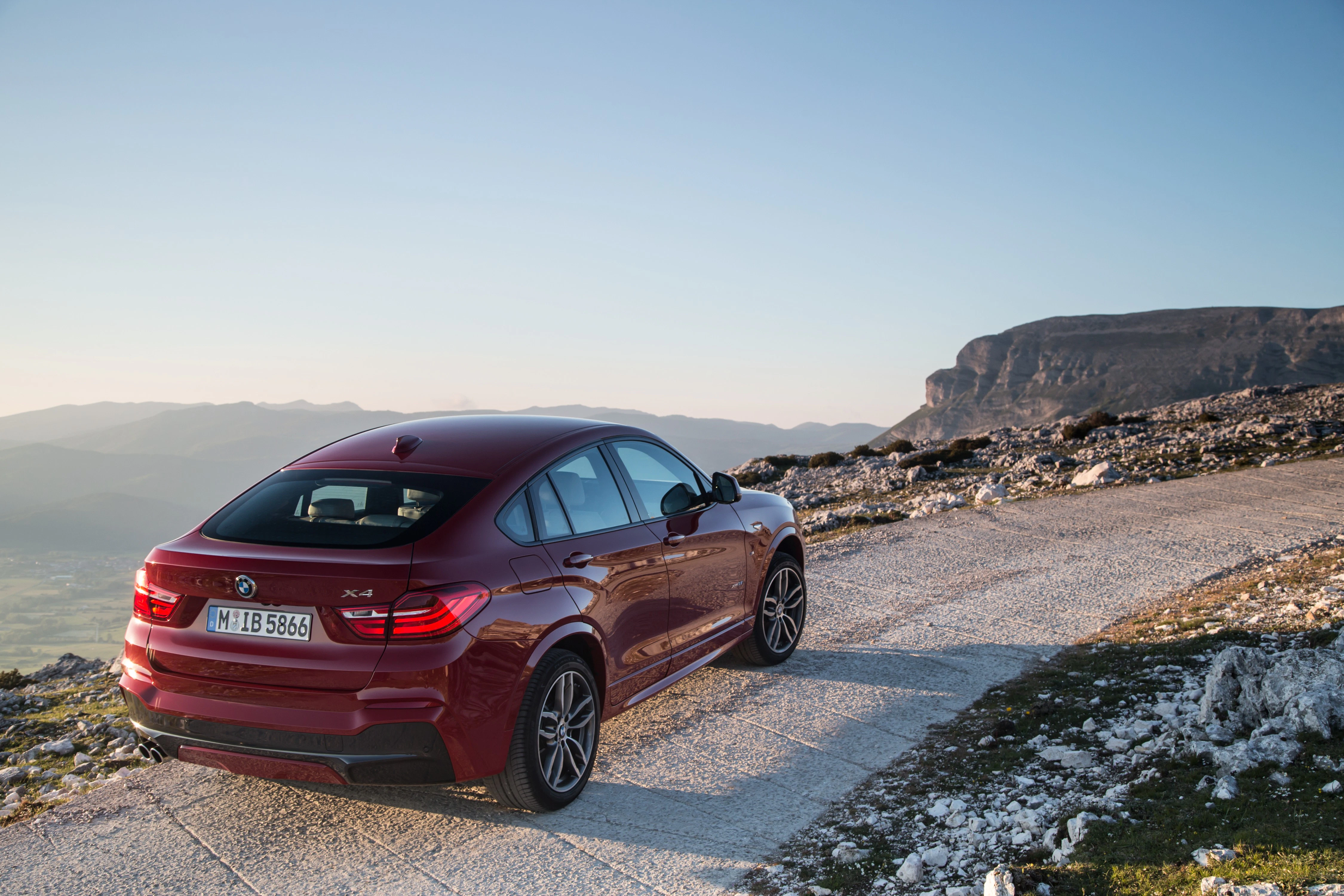 Roter SUV fährt auf einem schmalen, kurvigen Bergweg bei klarem Himmel und Blick auf ein weitläufiges Tal und Felsen.