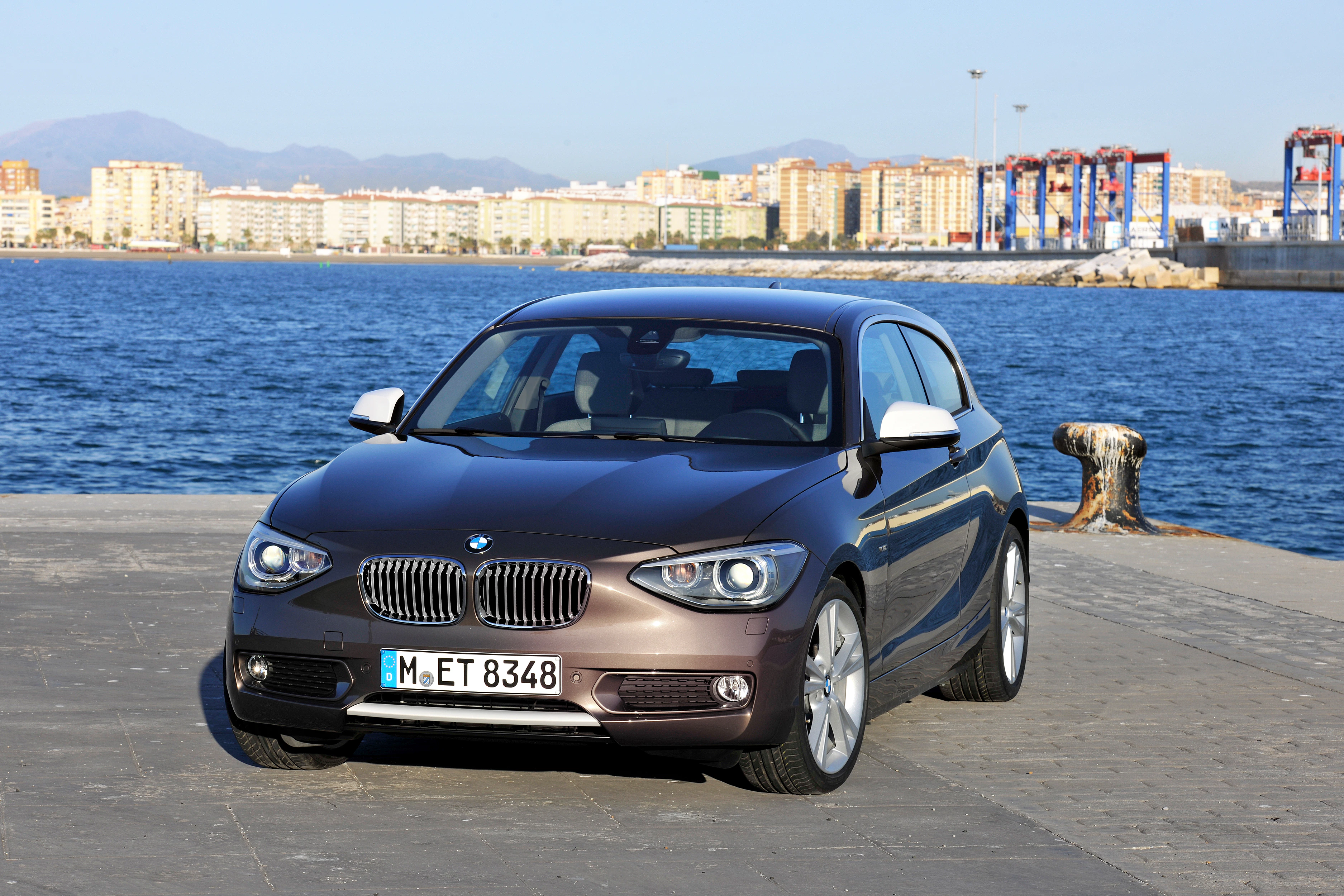 BMW 1er in Bronze steht auf einer Uferpromenade. Im Hintergrund das Meer und Gebäude in der Ferne. Klarer blauer Himmel.