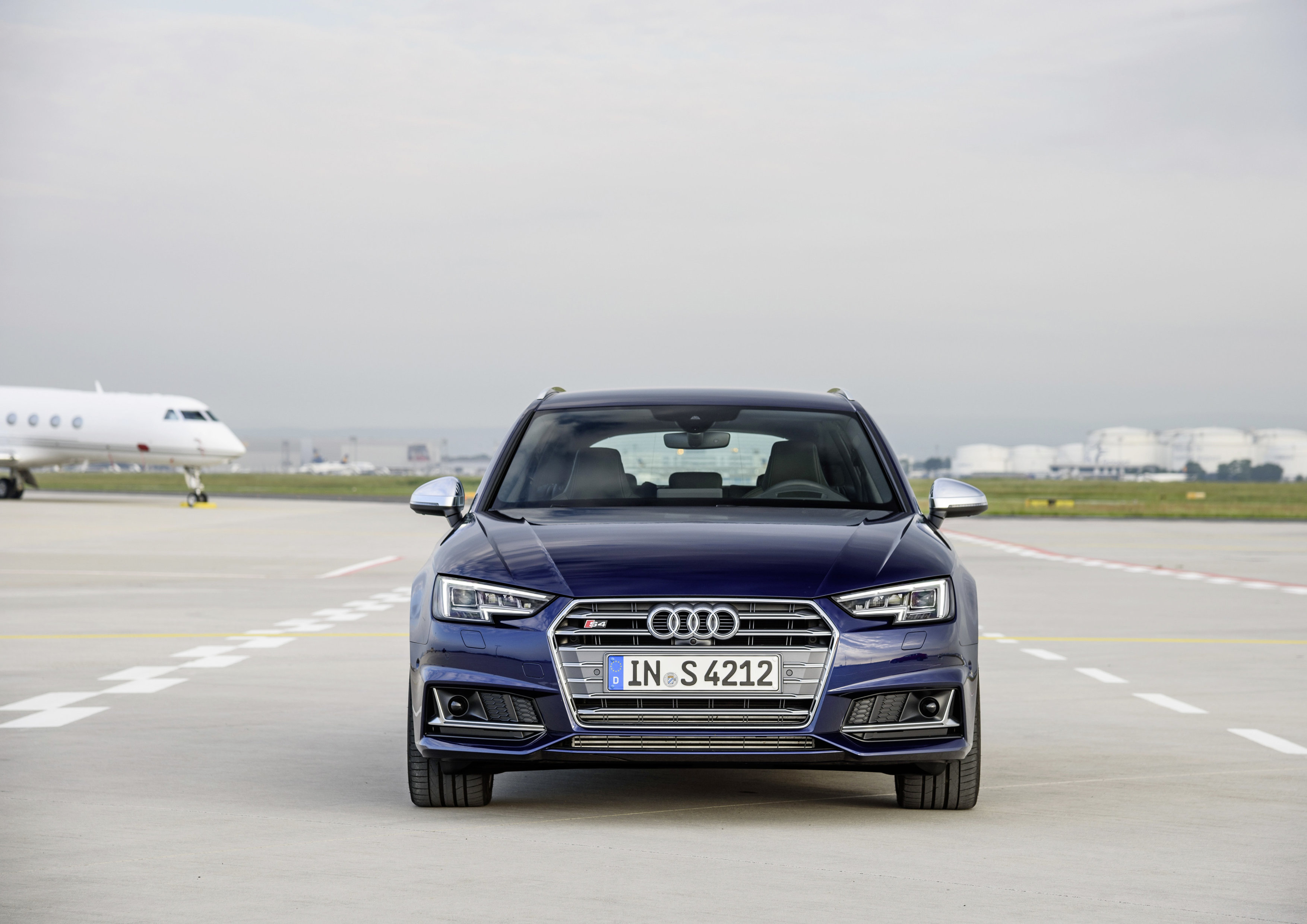 Blauer Audi, frontal aufgenommen auf einem Flughafen. Im Hintergrund sind ein startendes Flugzeug und Hangars zu sehen.