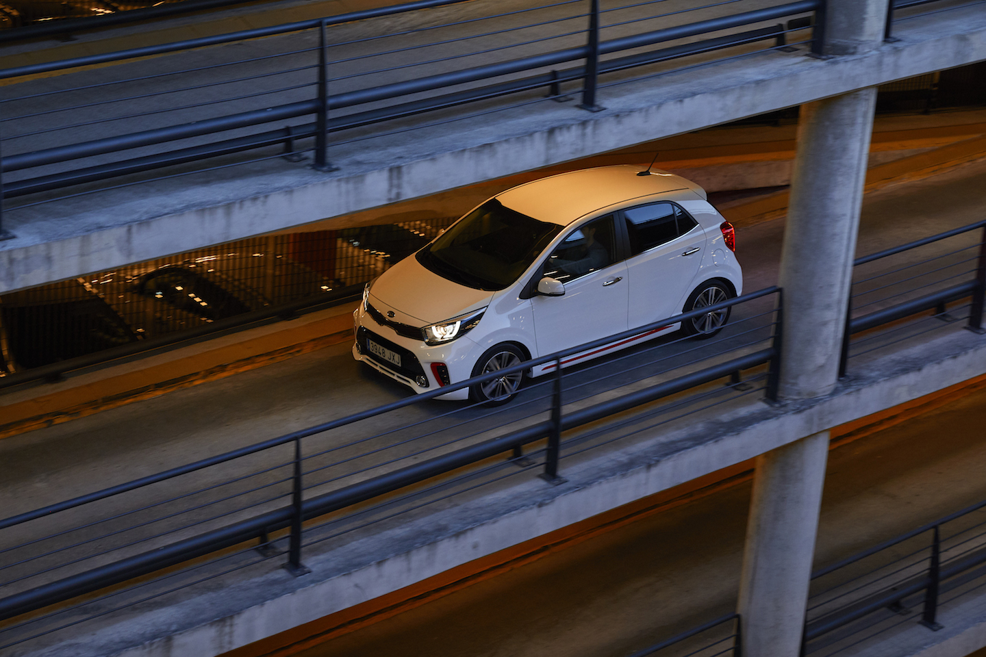 Weißes Auto fährt durch ein mehrstöckiges Parkhaus mit Betonwänden und Geländern. Subtile Beleuchtung umgibt das Fahrzeug.