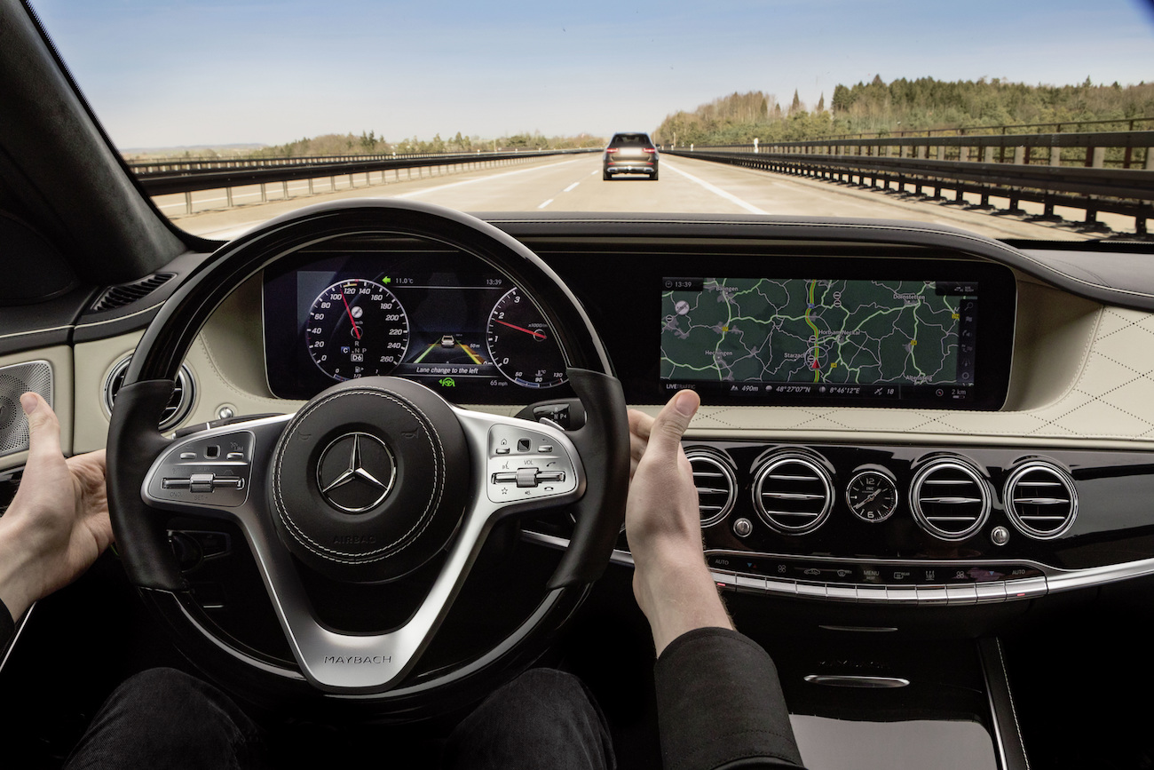 Person fährt Mercedes auf Autobahn, Lenkrad und Display zu sehen, Landschaft im Hintergrund.