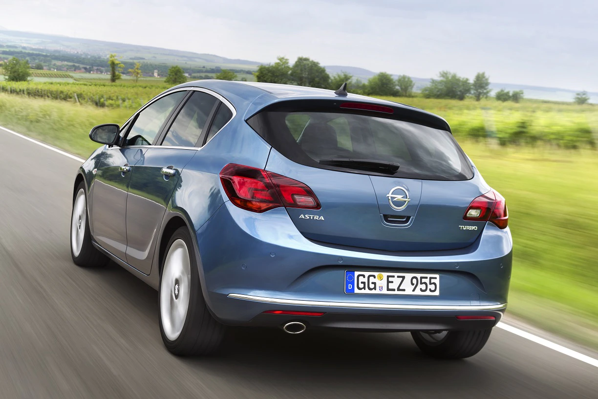 Ein blauer Opel Astra fährt auf einer Landstraße durch eine grüne Landschaft, bei bewölktem Himmel.