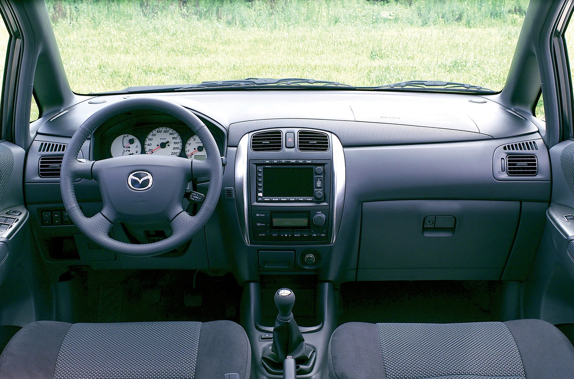 Innenraum eines Autos mit Mazda-Logo auf dem Lenkrad, zentralem Display und Schalthebel. Vordergrund mit zwei Sitzen, Hintergrund grüne Wiese.