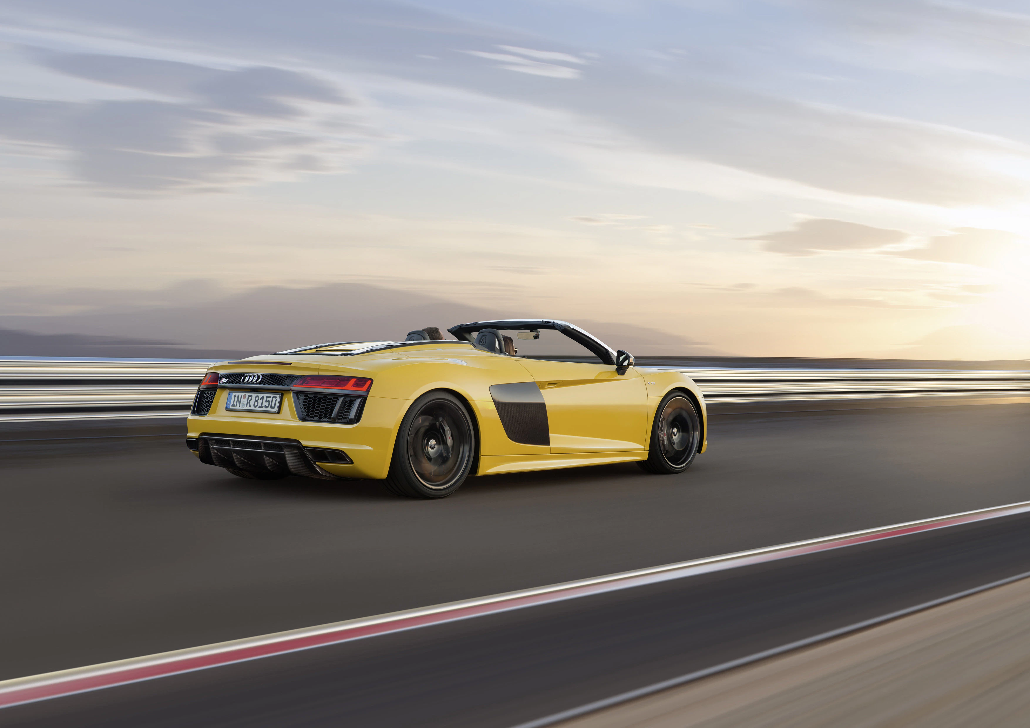 Gelber Audi R8 Spyder fährt schnell auf einer Straße mit verschwommenem Hintergrund bei Sonnenuntergang.