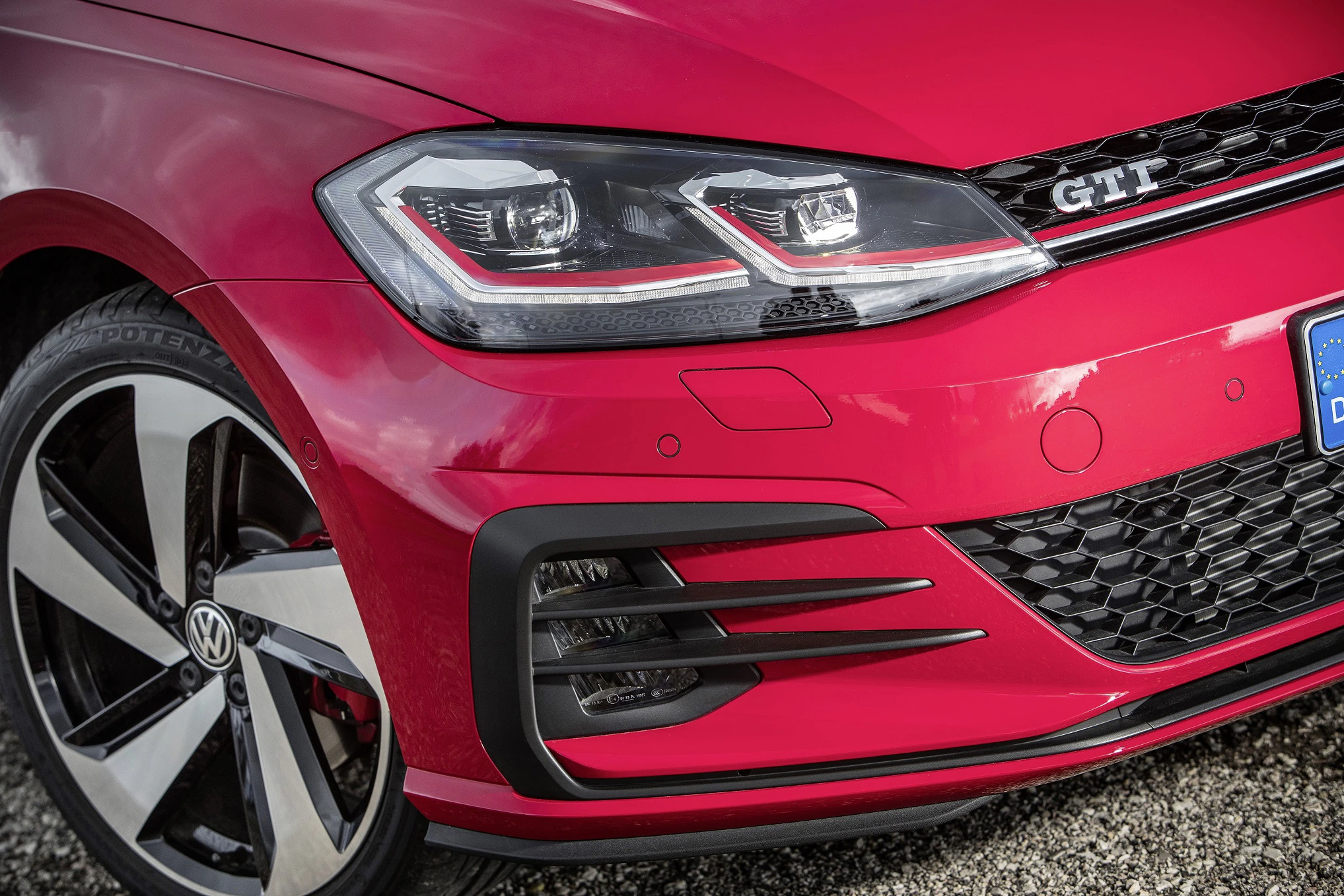 Nahaufnahme eines roten VW Golf GTI mit schwarzem Kühlergrill und auffälligen Scheinwerfern, abgebildet auf einer Kiesfläche.