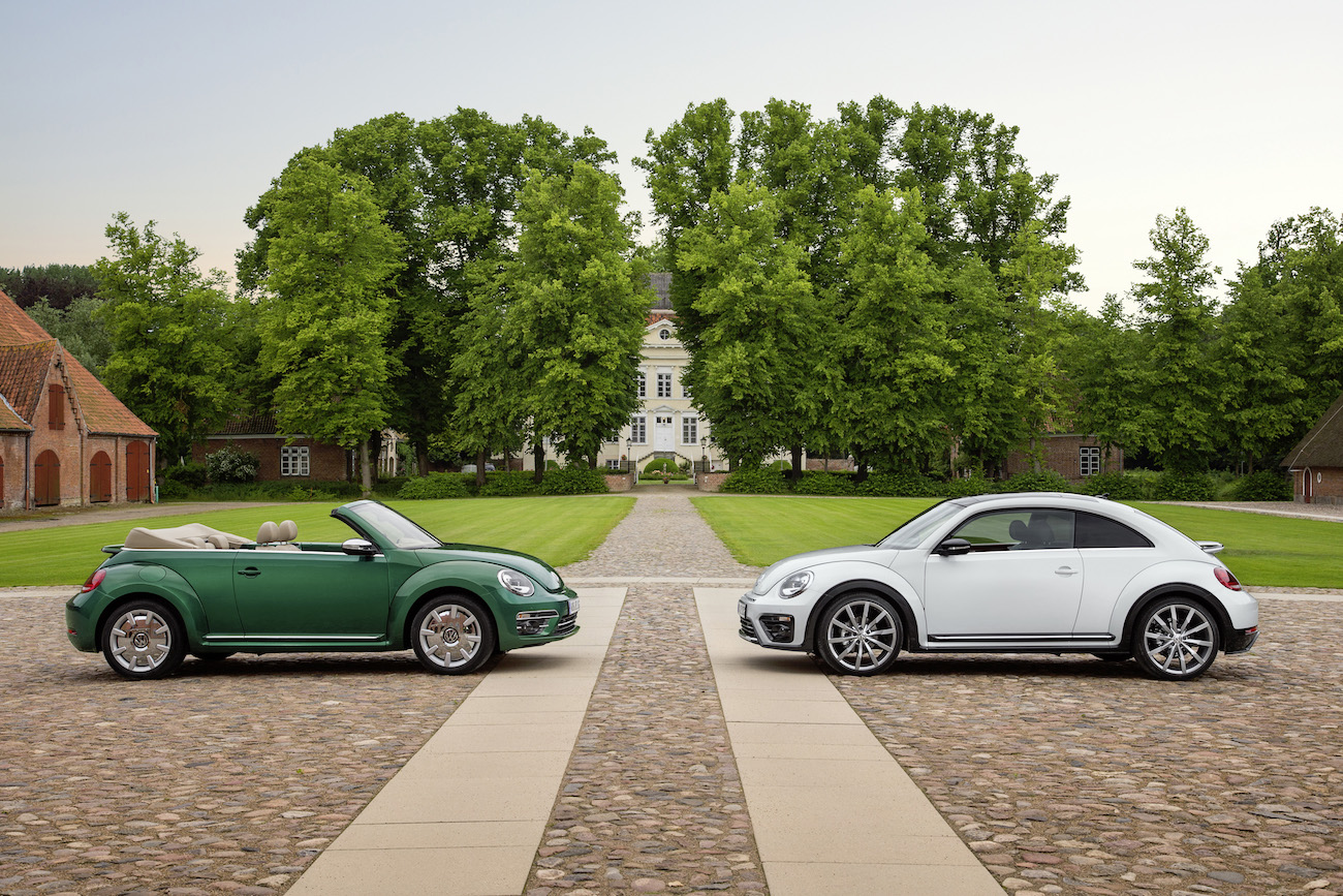 Zwei Volkswagen Beetle, grün und weiß, stehen auf gepflastertem Weg vor einem Herrenhaus mit grünen Bäumen im Hintergrund.