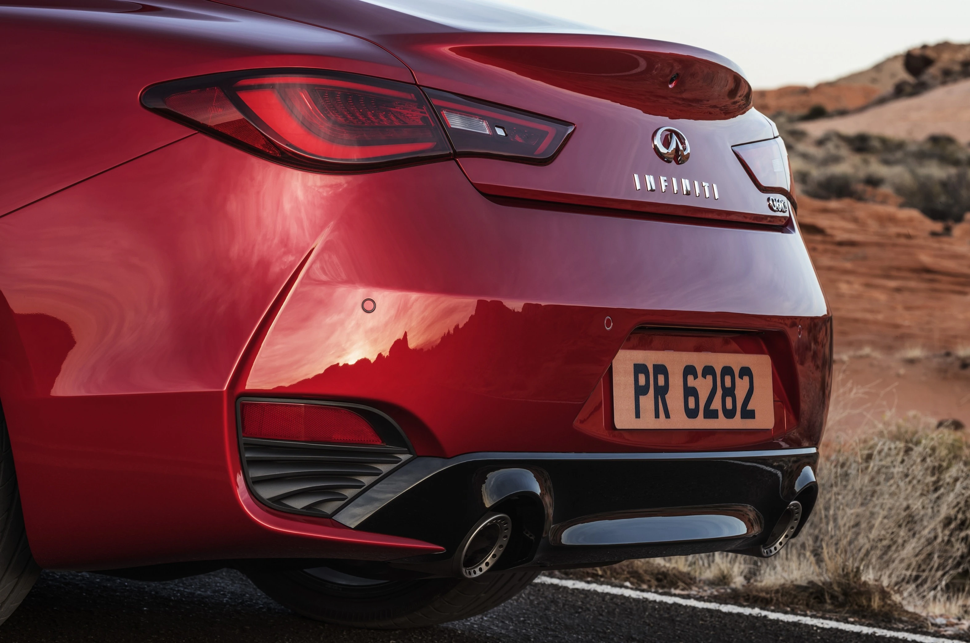 Rotes Auto von hinten fotografiert, Fokus auf Rücklicht, Auspuff und Nummernschild. Landschaft mit Hügeln im Hintergrund.