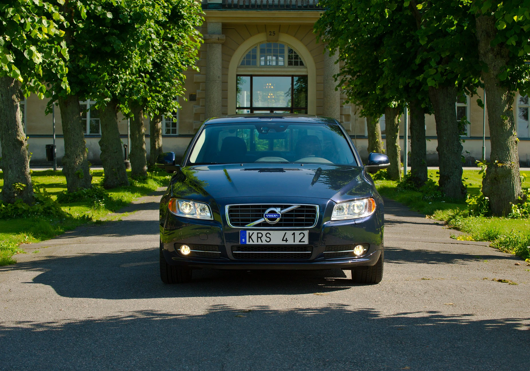 Ein schwarzer Volvo steht frontal auf einer von Bäumen gesäumten Auffahrt vor einem großen Gebäude.