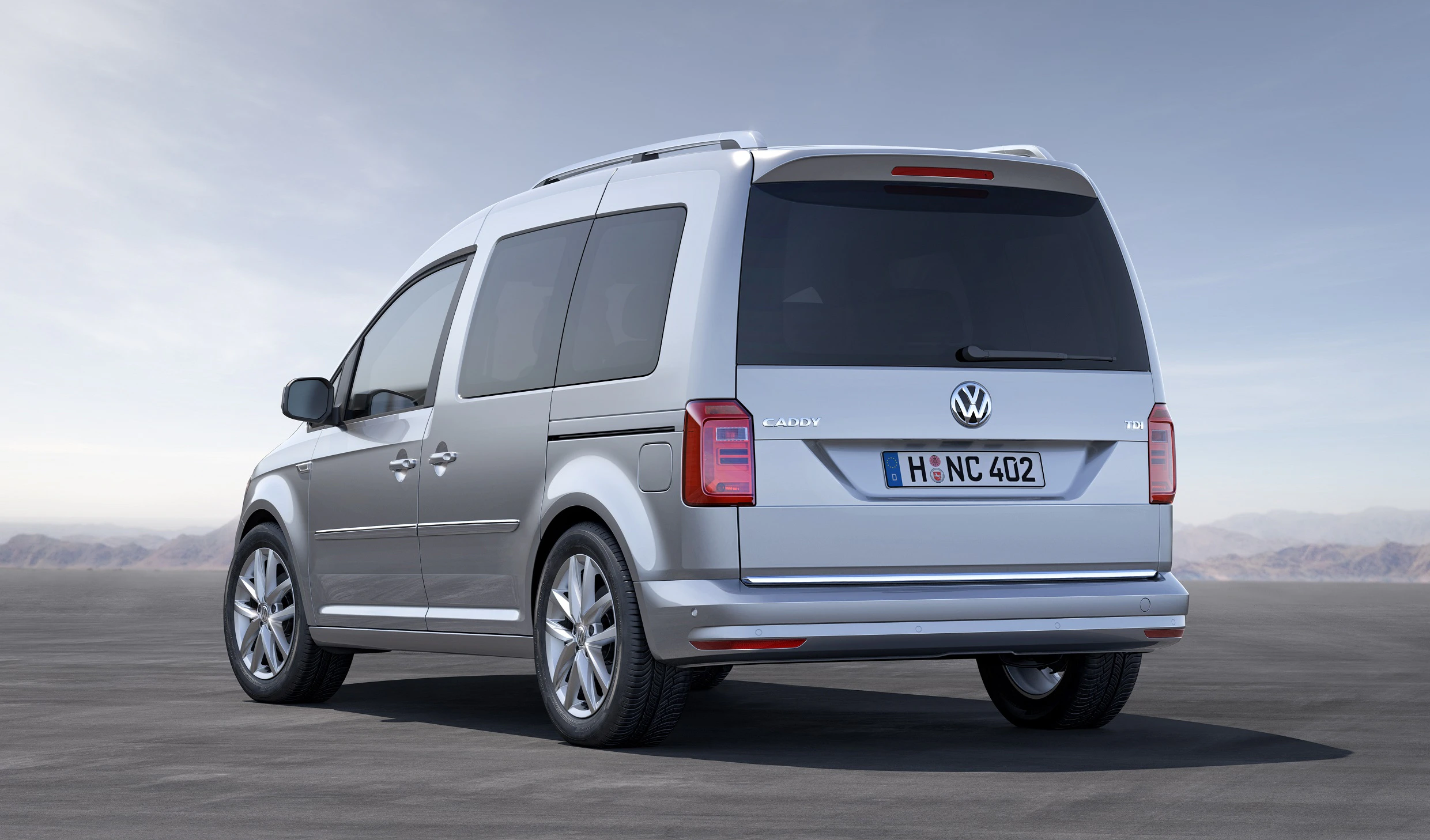 Silberner VW Caddy auf asphaltierter Fläche, Rückansicht. Im Hintergrund ein klarer Himmel und Berge in der Ferne.