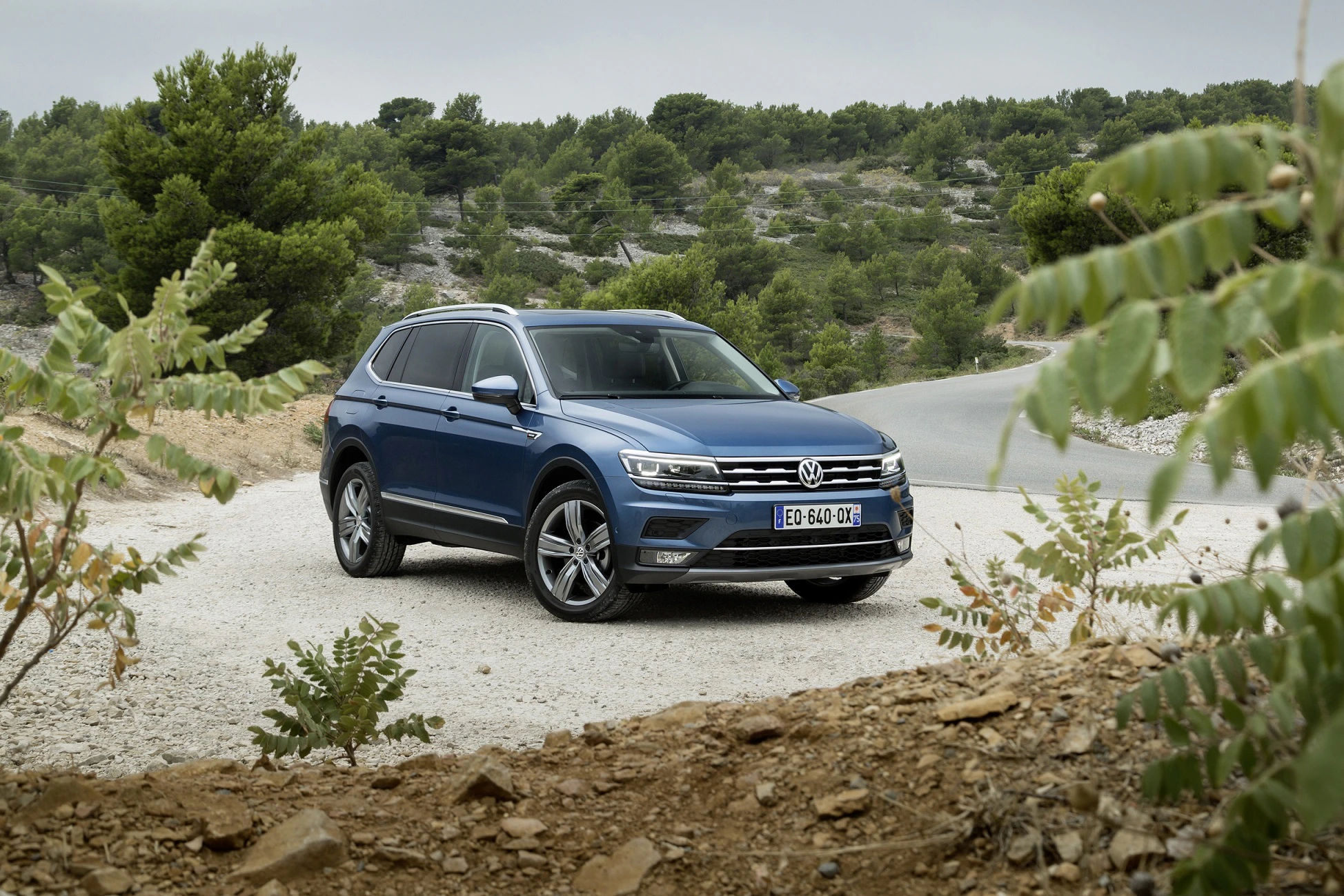 Blauer SUV fährt auf Schotterstraße durch eine hügelige, bewaldete Landschaft. Auto ist von einer Biegung der Straße umgeben.
