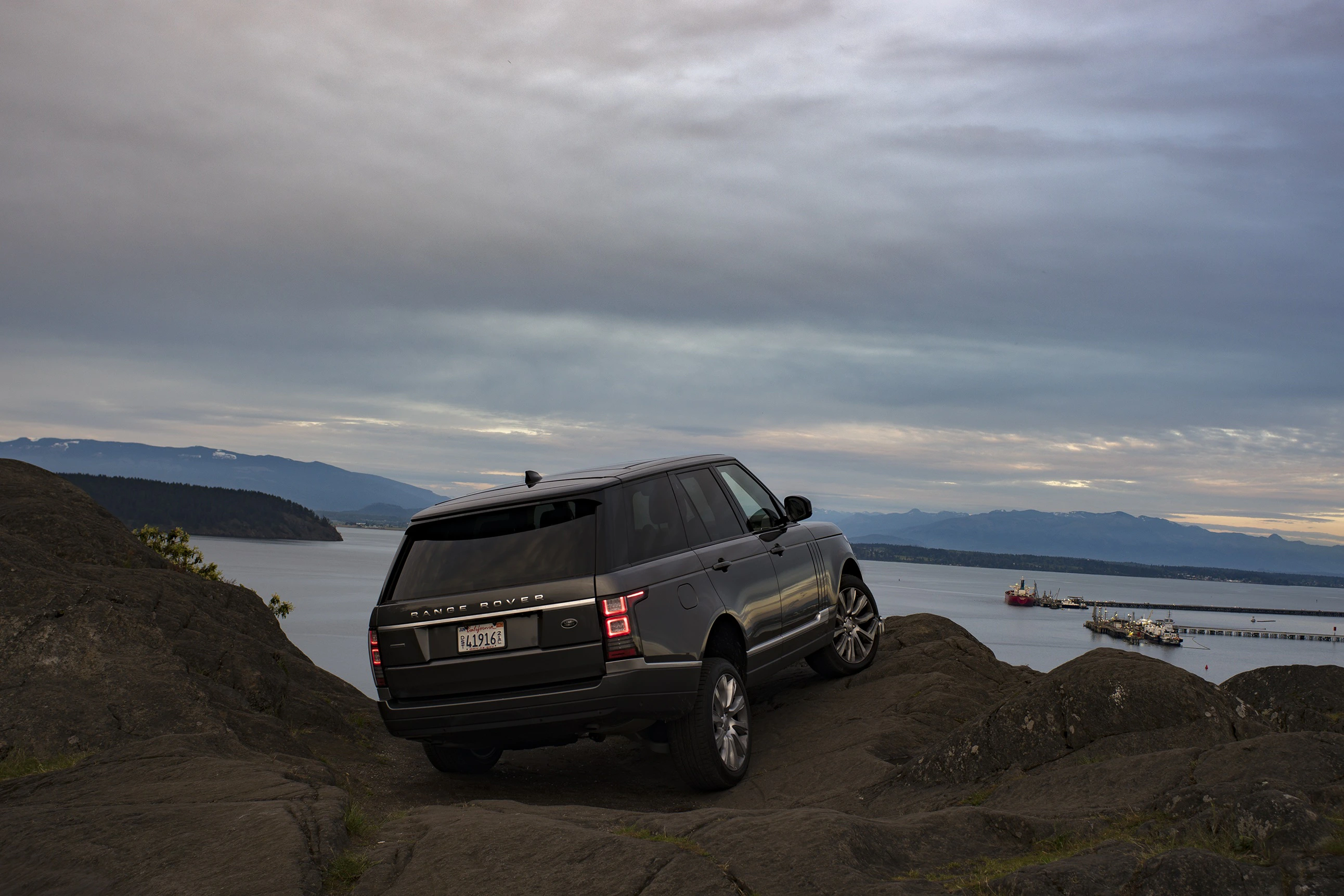 Ein schwarzer SUV fährt über eine felsige Klippe mit Blick auf ein Gewässer und bewölkten Himmel im Hintergrund.