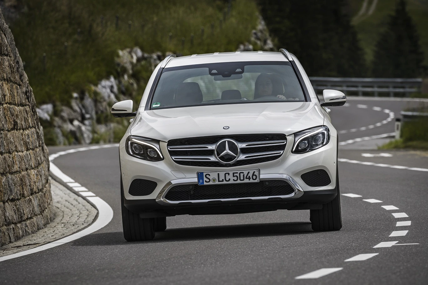 Weißer Mercedes-Benz SUV fährt auf kurviger Bergstraße, gesäumt von Steinmauer und grüner Landschaft.