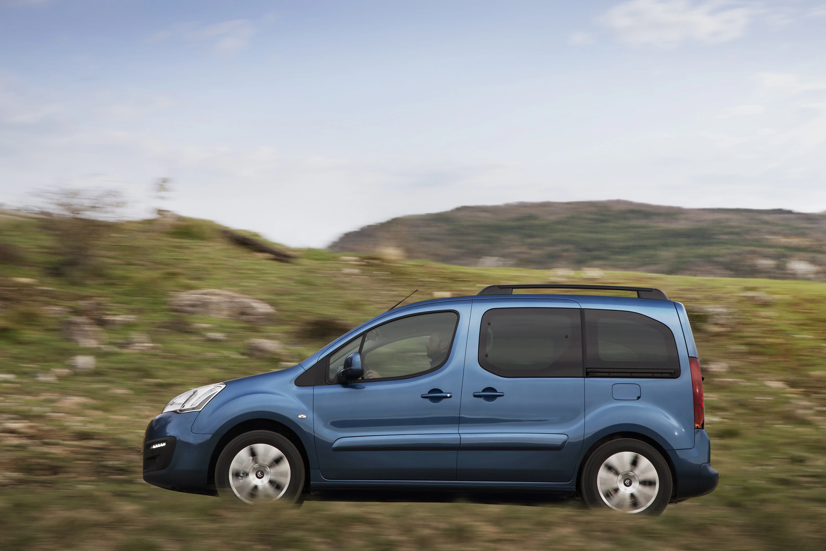 Blauer Minivan fährt auf einer grünen Wiese mit bewölktem Himmel und Hügeln im Hintergrund.