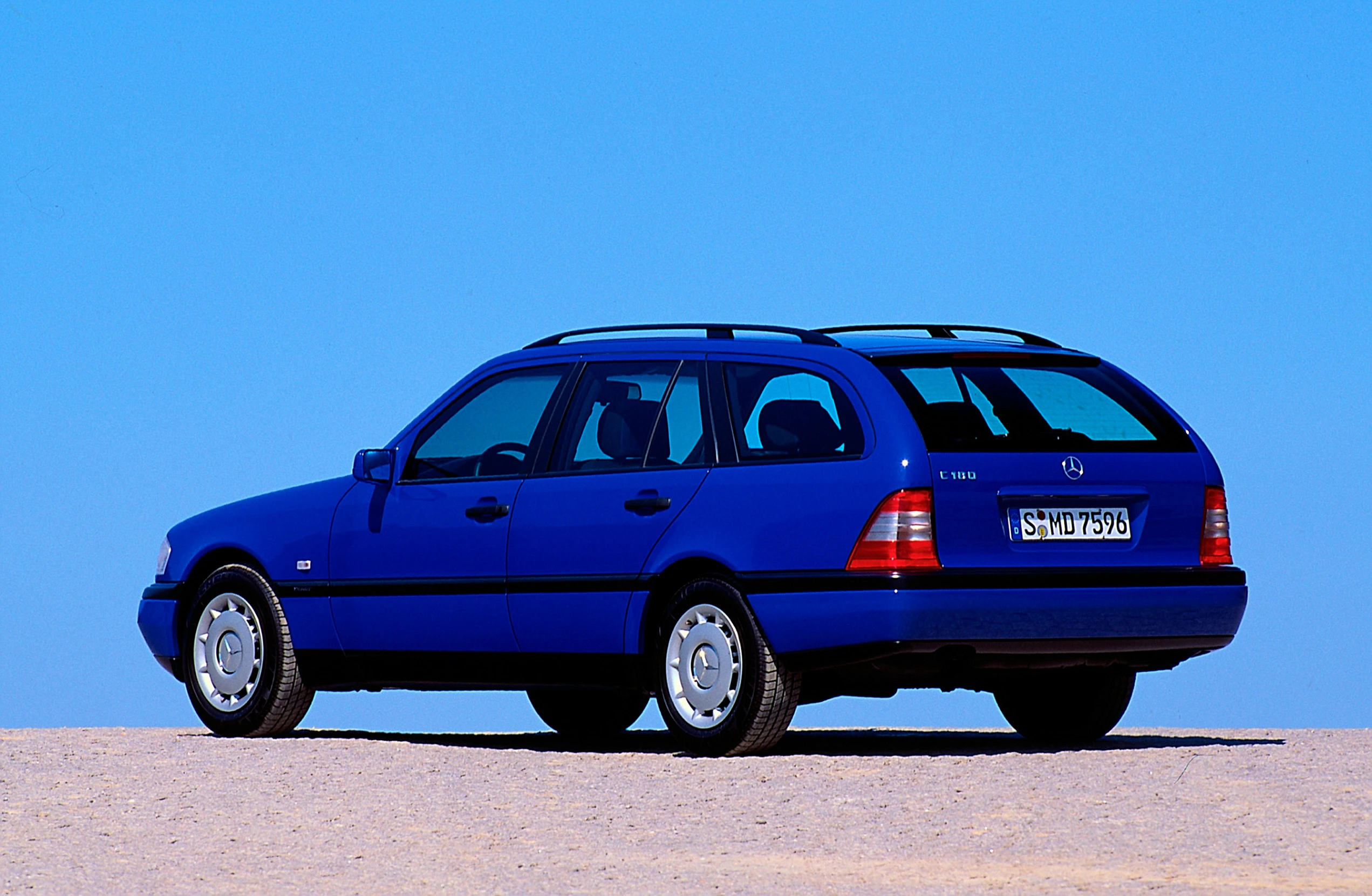 Blauer Kombi auf Sandboden, seitlich von hinten fotografiert vor klarem Himmel, Kennzeichen sichtbar. 