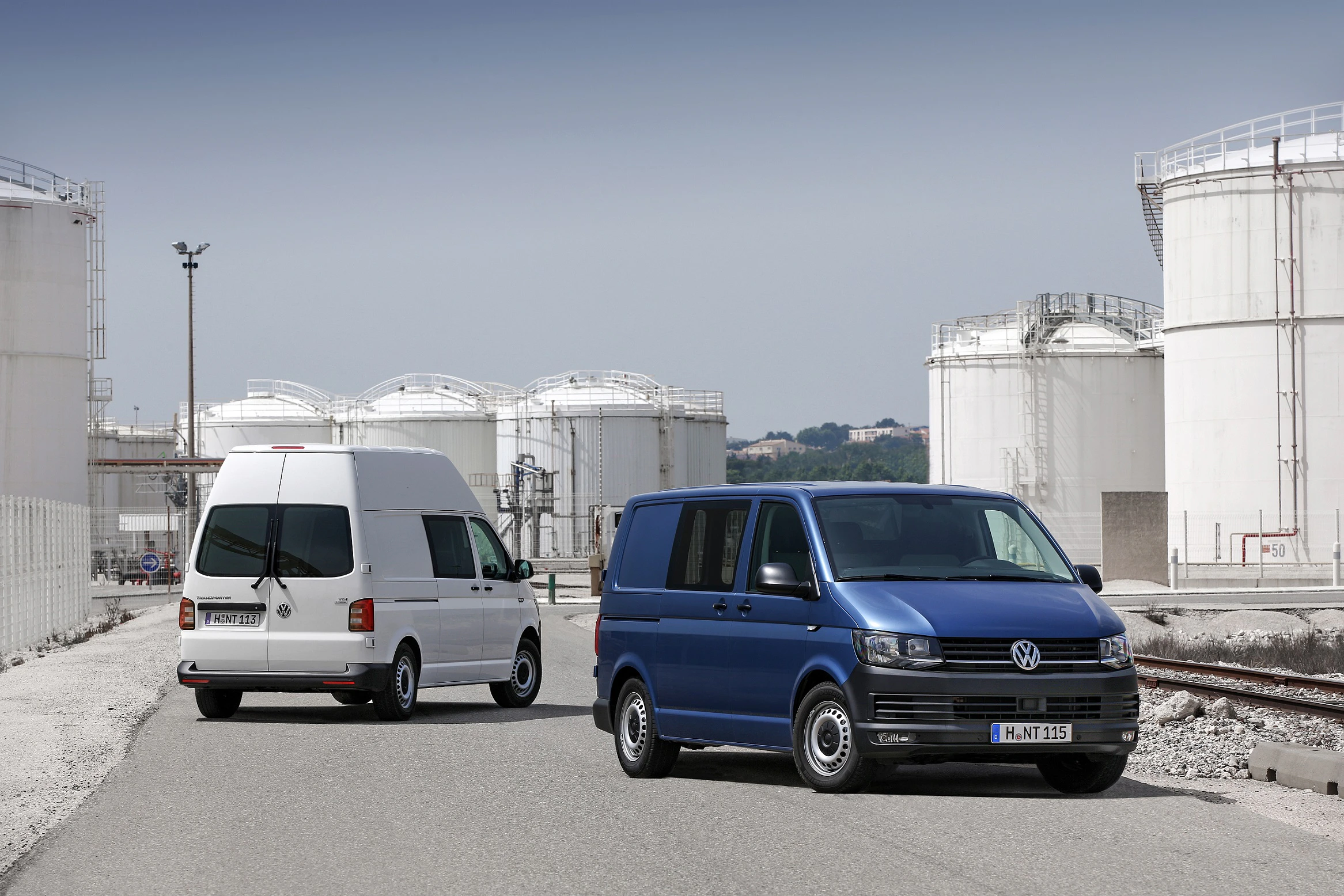 Zwei VW-Transporter parken vor großen, weißen Industrieanlagen. Vordergrund blau, Hintergrund weiß. Klarer Himmel.